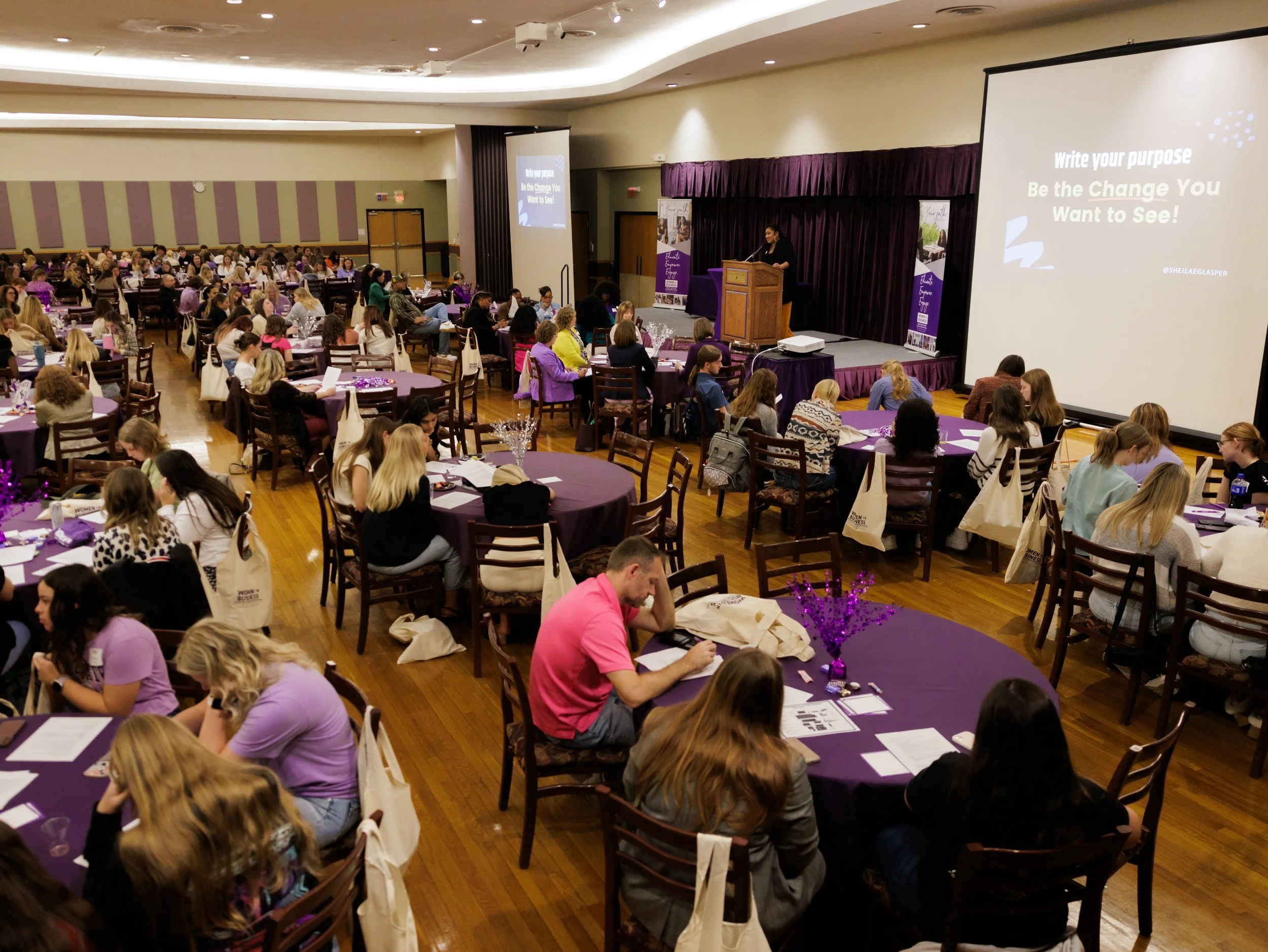 Large conference room filled with people seated at round tables, a stage with a screen and purple curtains at the front.