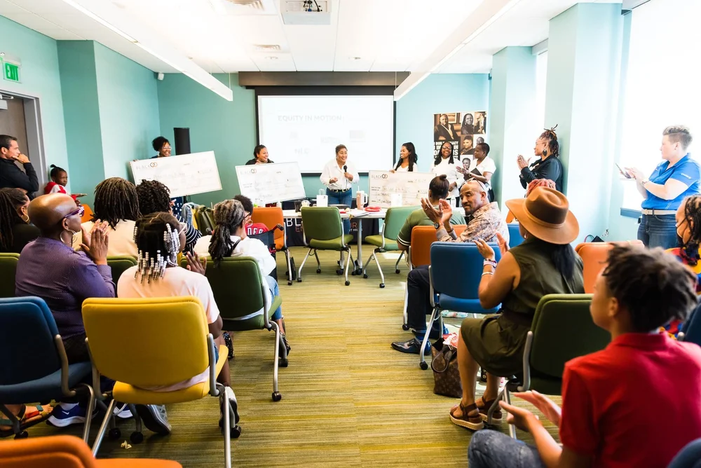 People seated in a room attending a panel discussion with a speaker at the front, large screen, and photos on the wall.