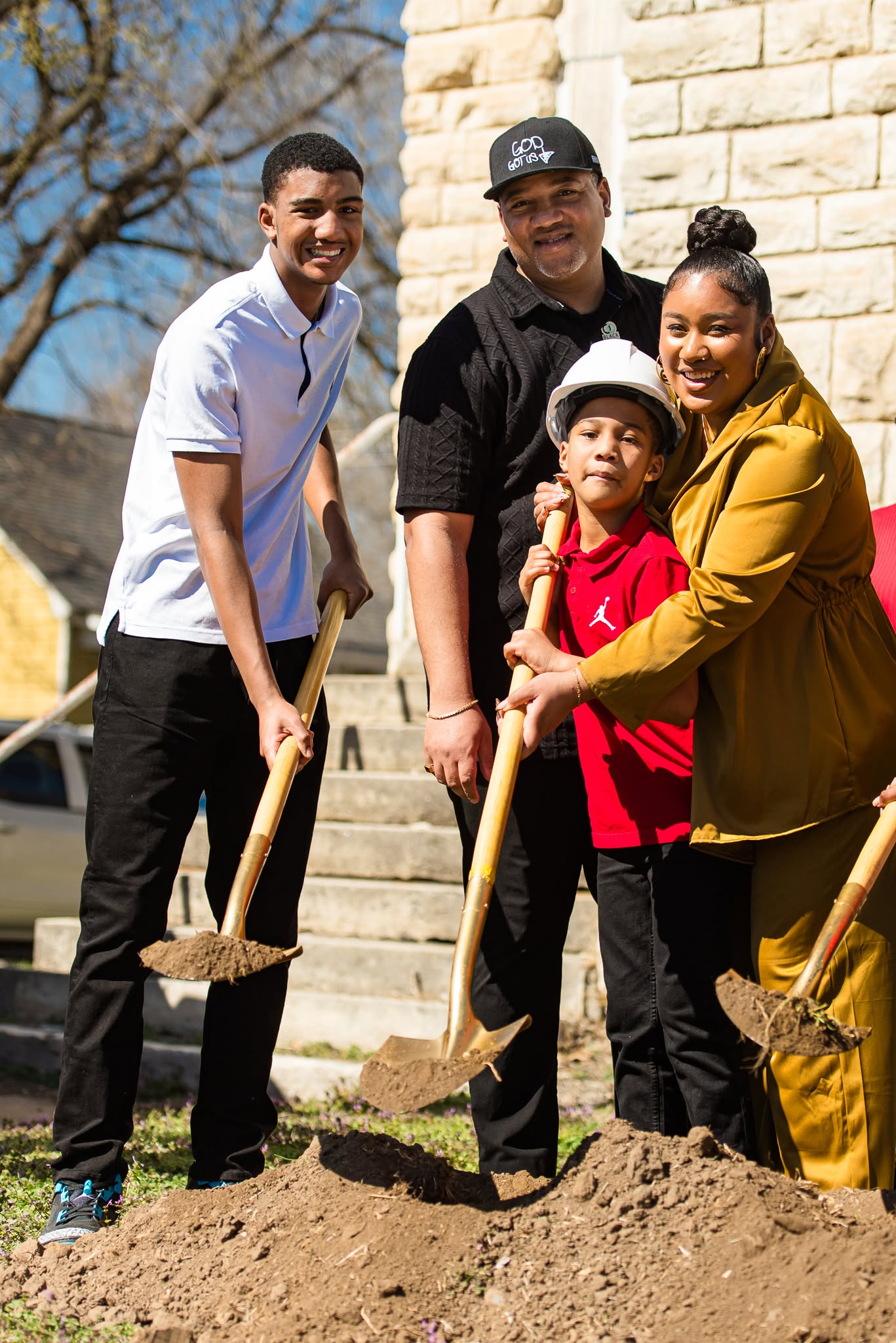 Group of four people with shovels outdoors, smiling, standing in front of a stone wall, planting or digging in soil.