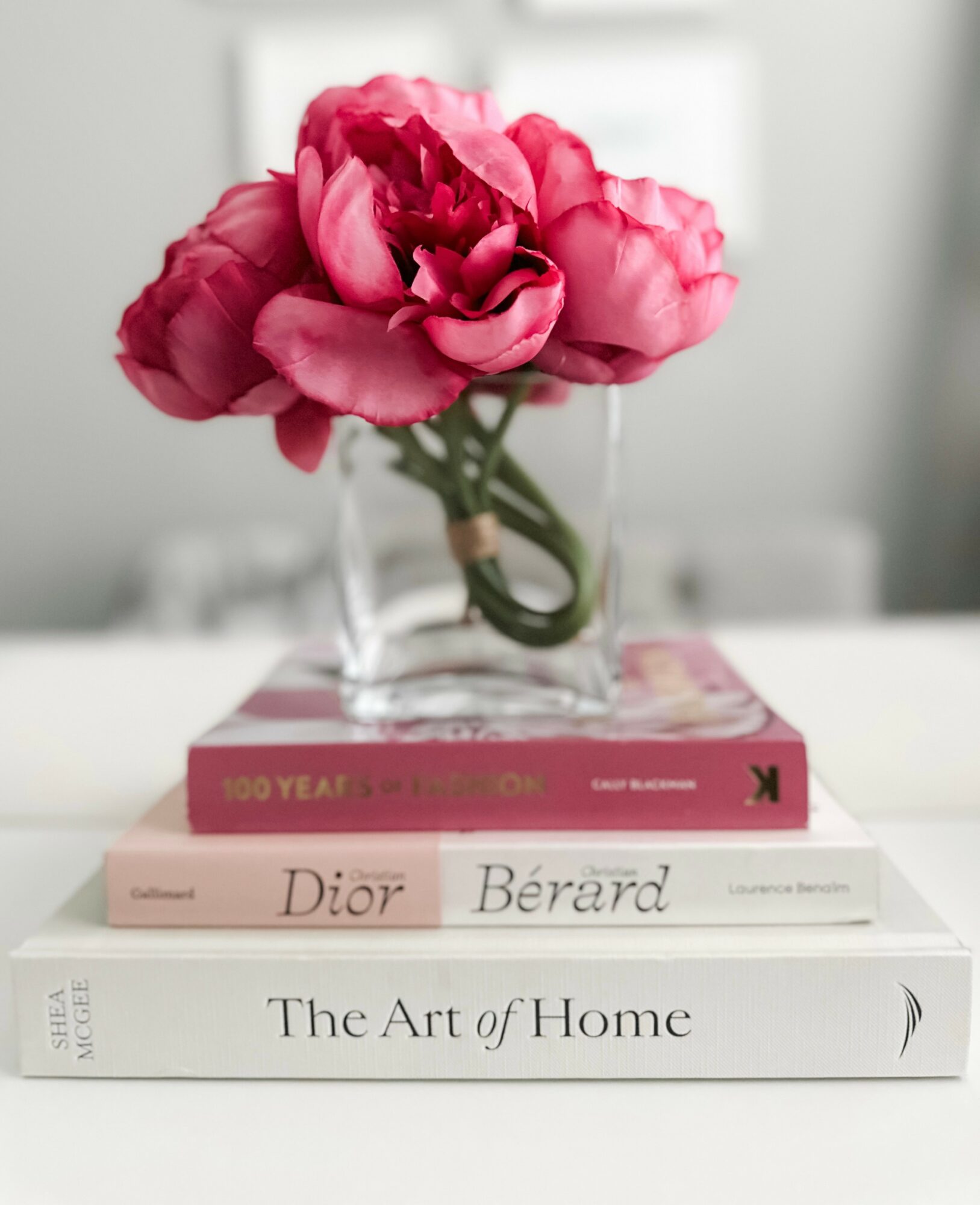 Pink peony in a glass vase on a stack of books on a white table, blurred background, neutral tones.