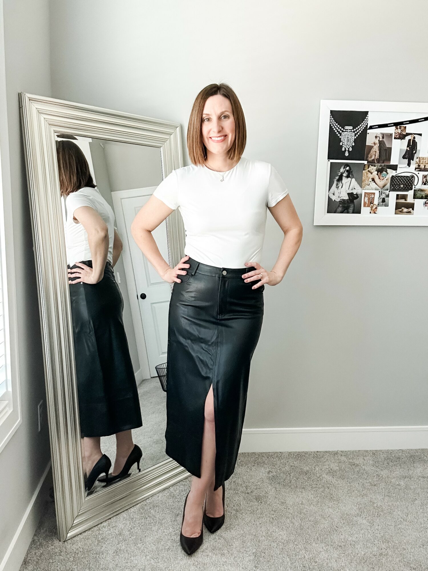 Woman with shoulder-length hair in a white top and black skirt standing in front of a mirror, smiling, with a collage on the wall behind her.