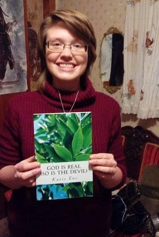 Person with glasses and short hair holding a book with green leaves on the cover, smiling indoors.