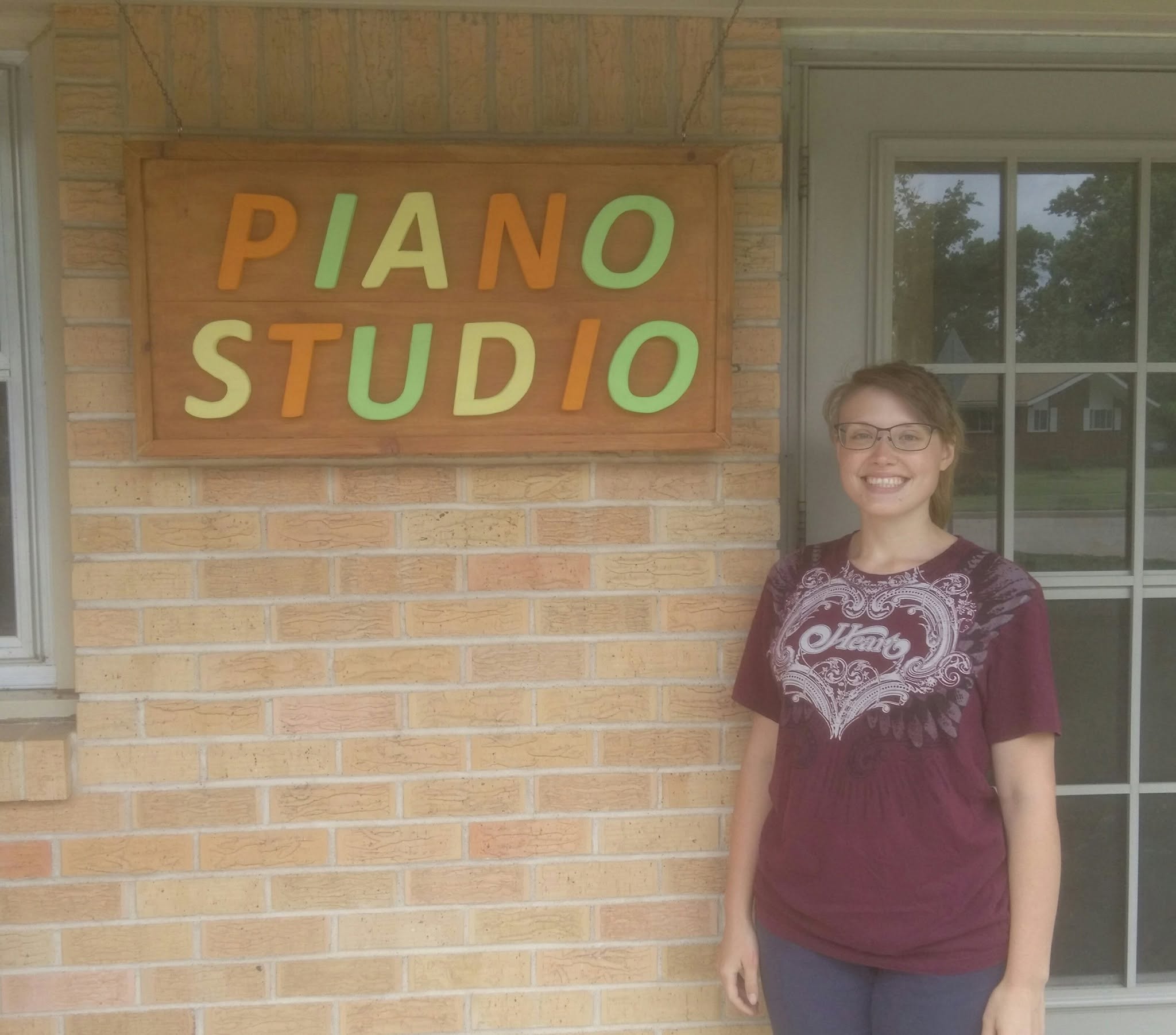 Girl standing outside next to a sign that reads 'PIANO STUDIO' on a brick wall, smiling, wearing glasses and a maroon shirt.