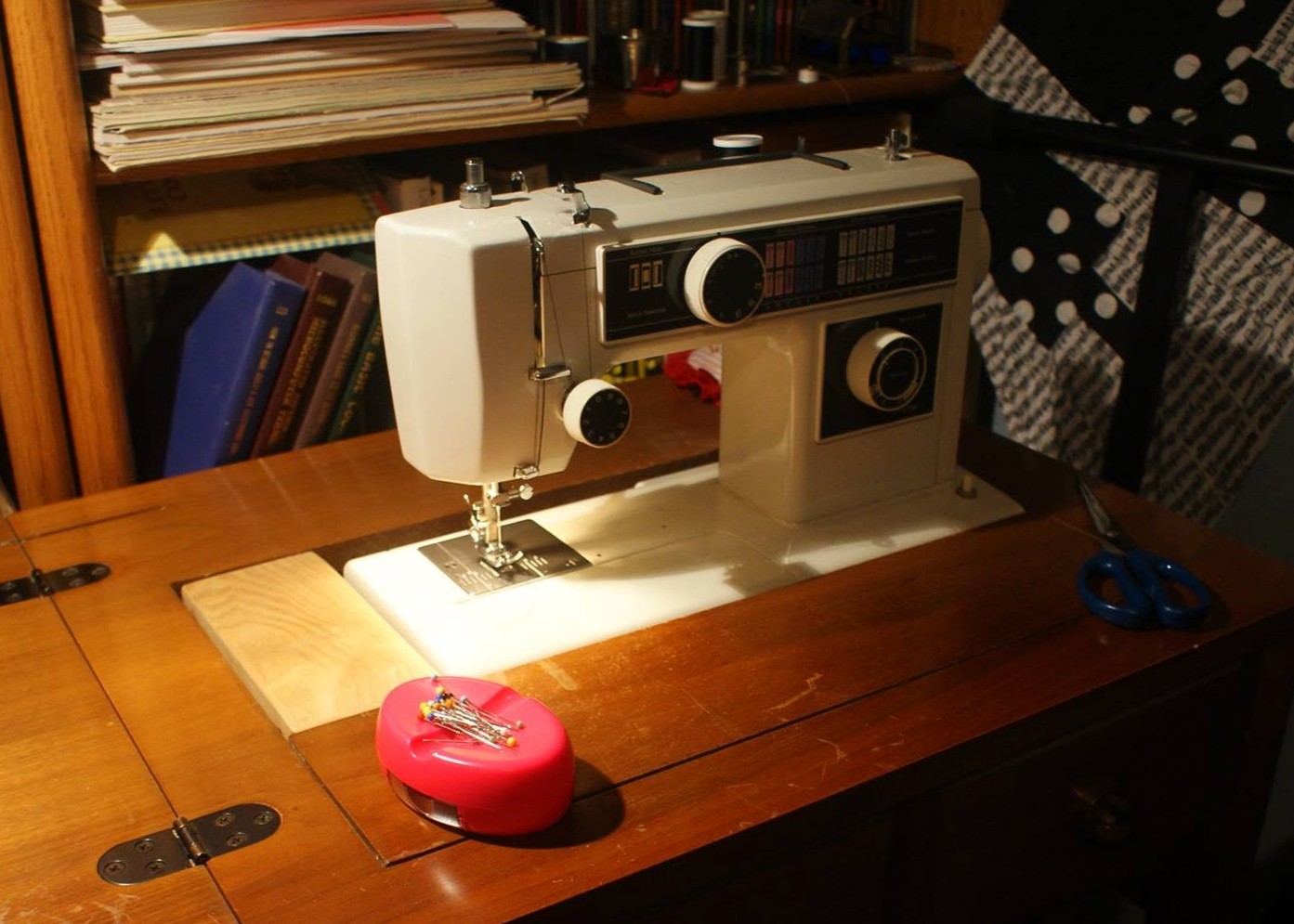 Sewing machine on wooden table with pink tape measure and scissors nearby, books on shelf in background.