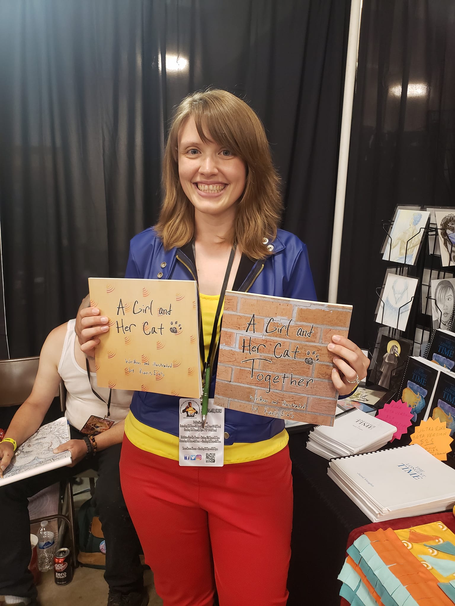 Woman smiling holding two handmade signs at an event booth, with black curtains and display materials in background.