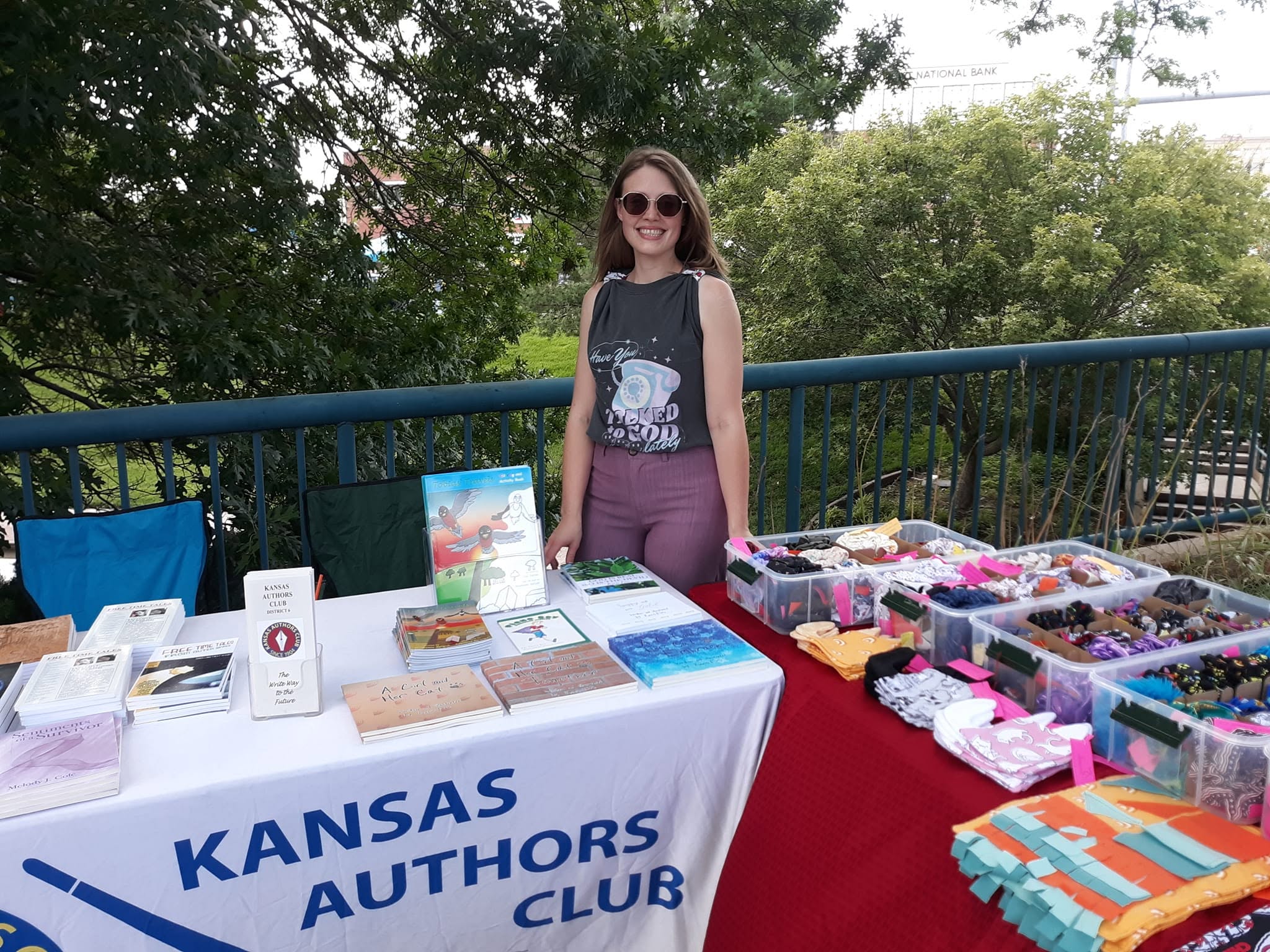 Woman standing behind a table with books and jewelry outdoors, smiling, trees in background.