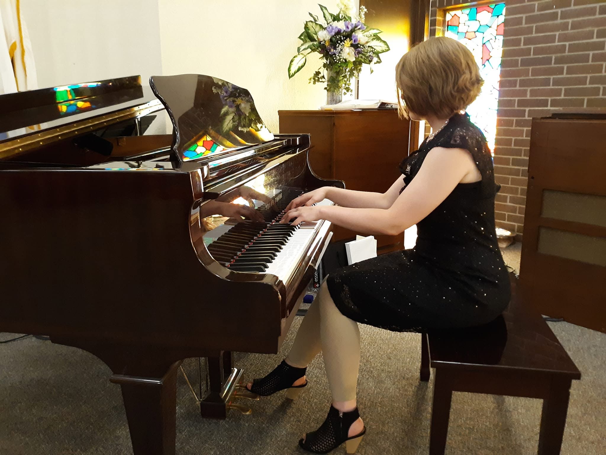 Child playing piano in a room with a flower arrangement and brick wall, wearing a black dress and heels.