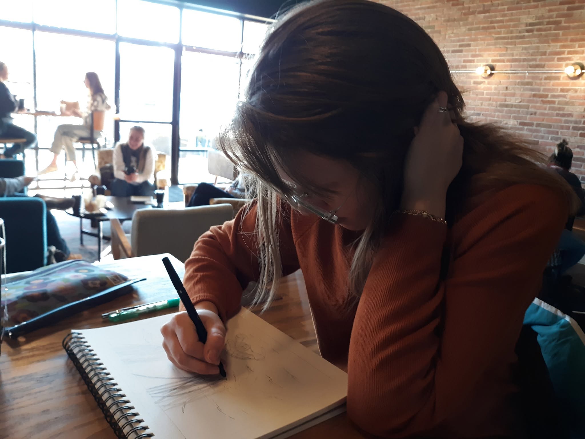 Person with long hair writing in a notebook at a table in a cafe, with people and large windows in the background.