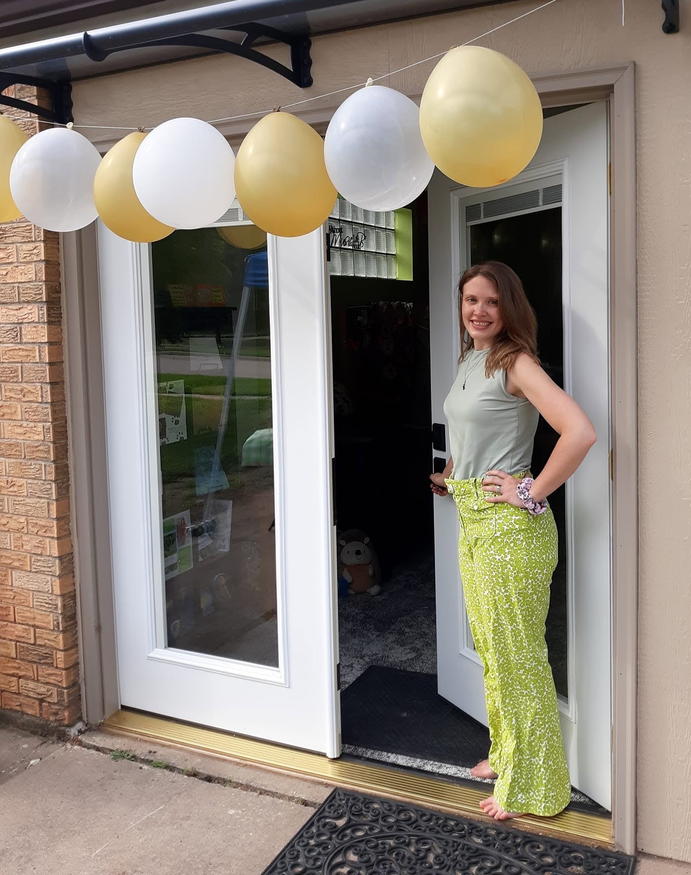 Woman standing in doorway with balloons overhead, smiling, wearing a sleeveless top and bright yellow pants, holding a drink.