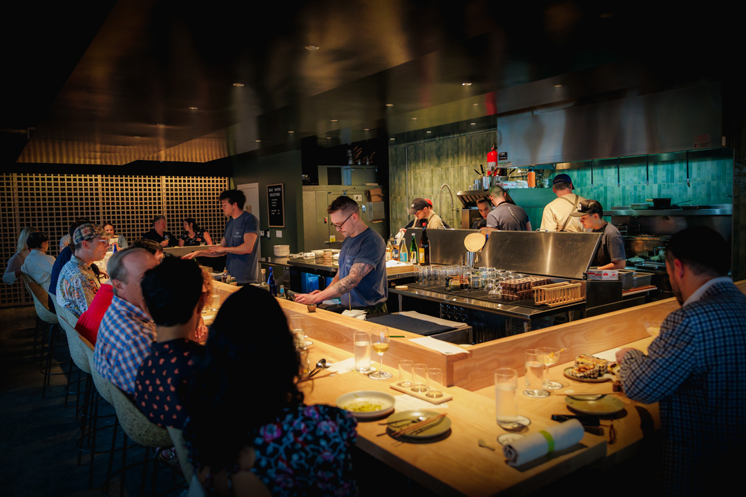 People sit at a bar counter watching chefs prepare food in a restaurant kitchen, with a dark background and warm lighting.