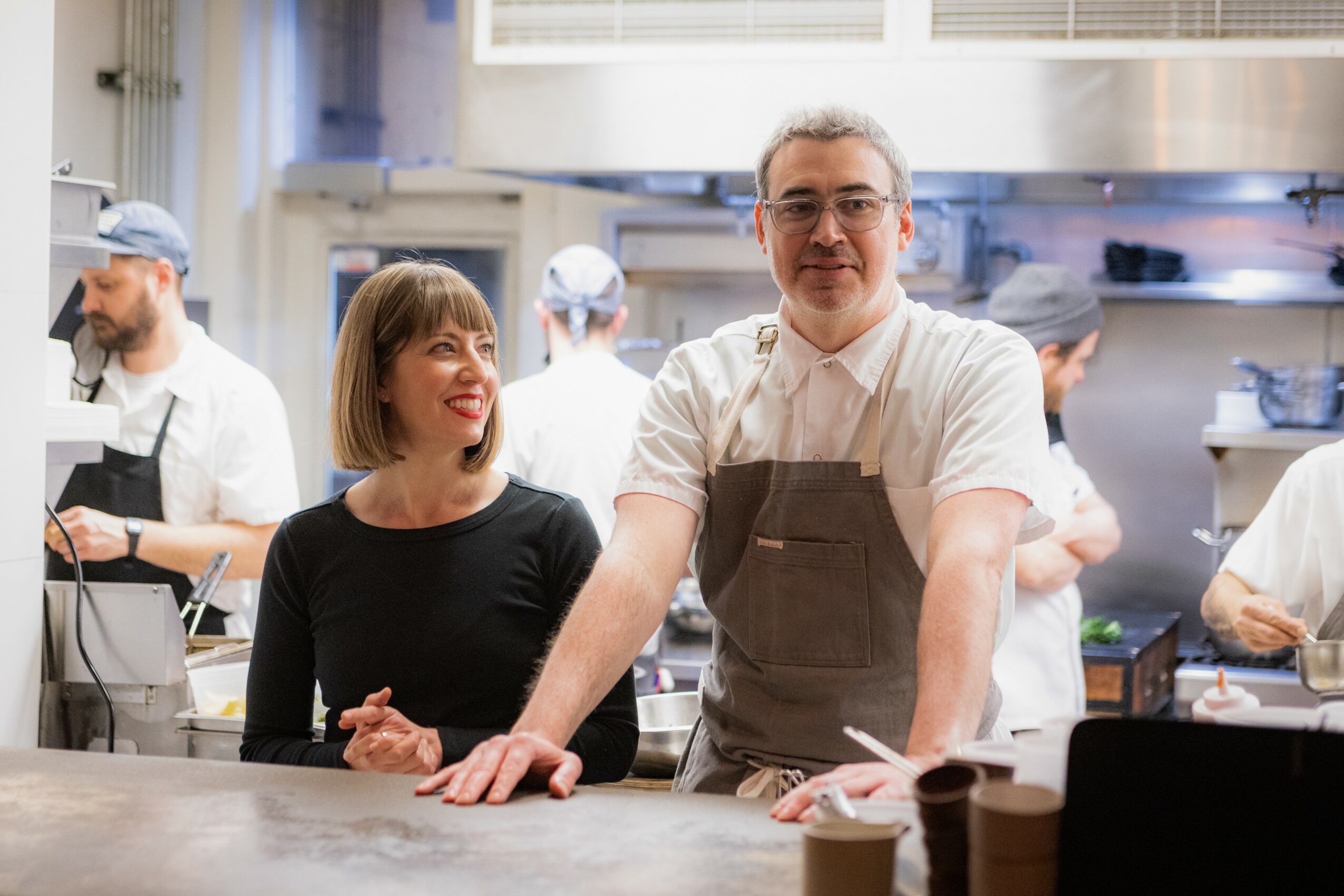 Chef and woman smiling in a busy kitchen, with other staff working in the background.