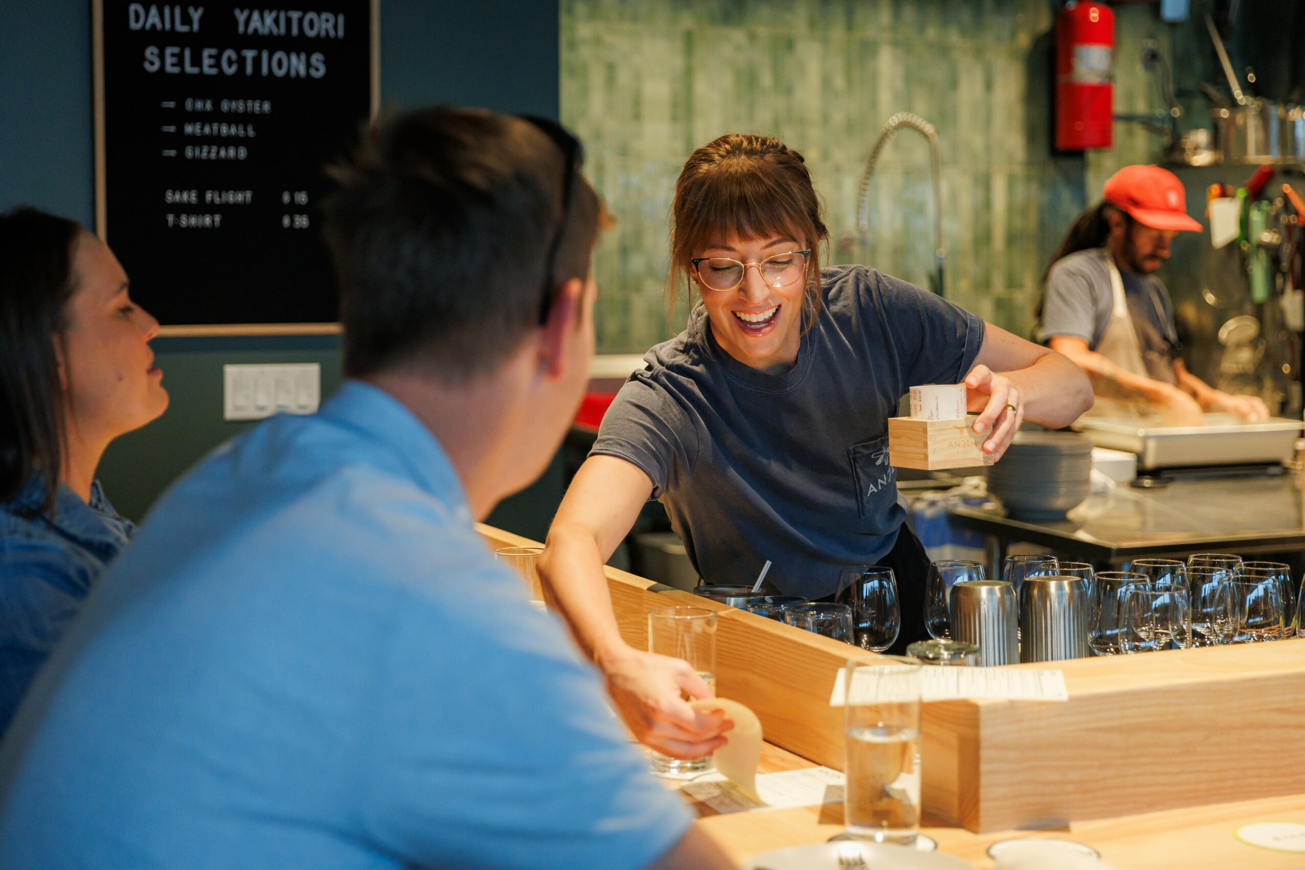 A smiling woman serving drinks to two customers at a bar counter in a lively restaurant or bar setting.
