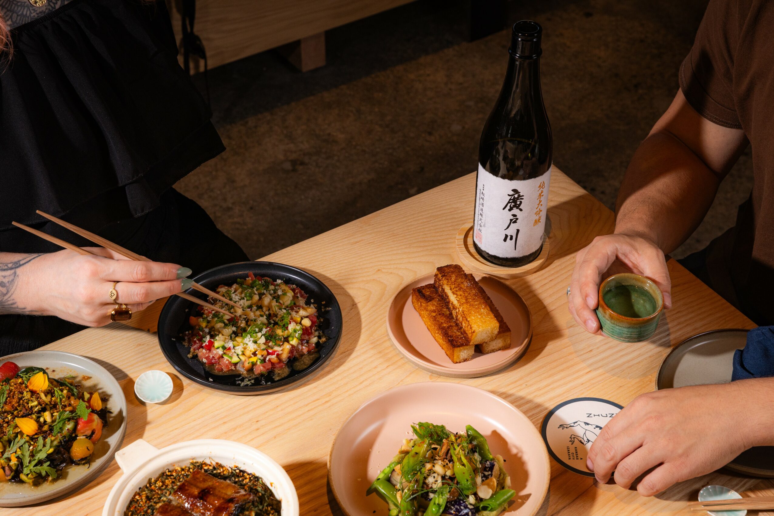 People dining with dishes, a bottle, and a drink on a wooden table, with hands reaching for food and utensils.