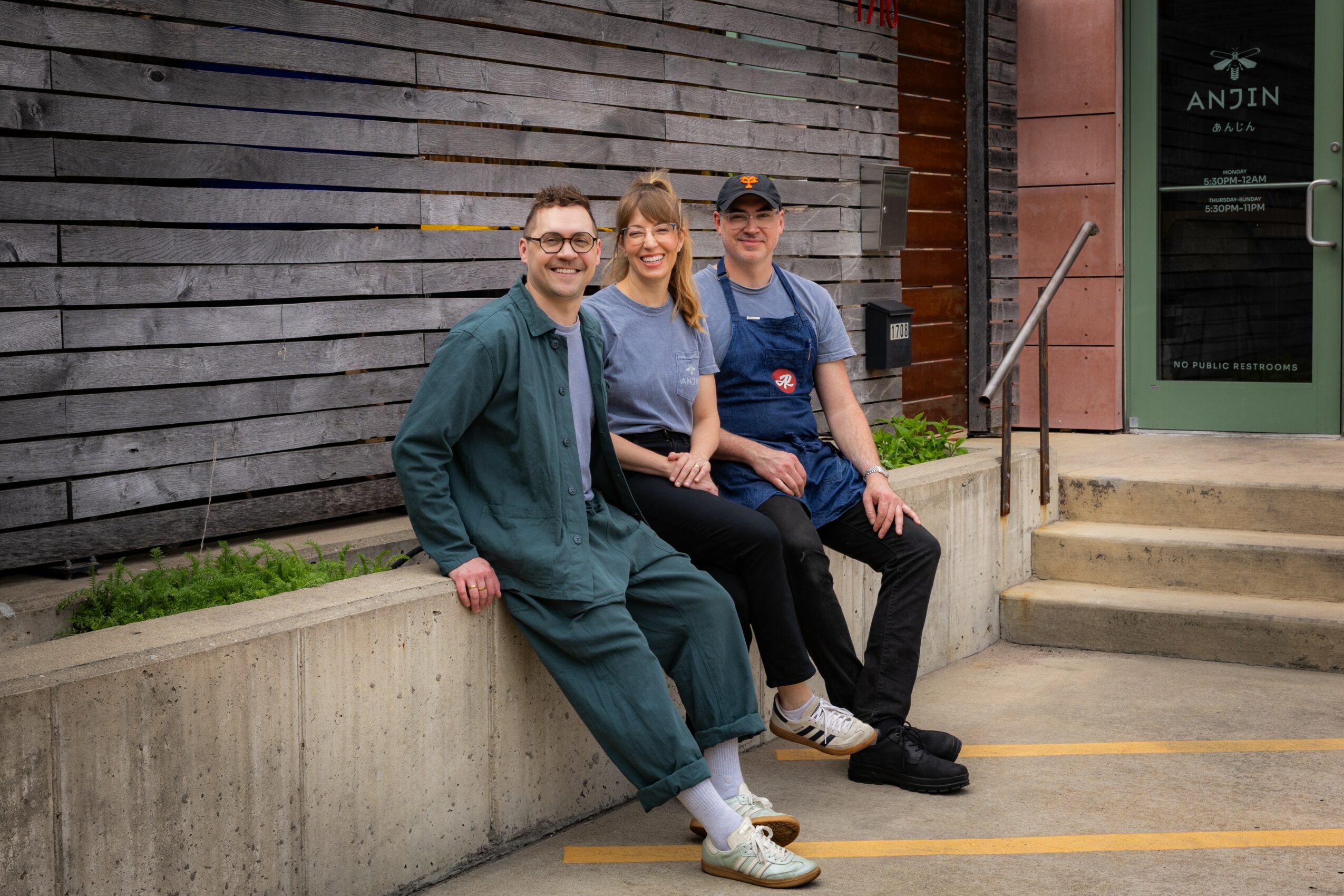 Three people sitting on a concrete ledge outside a building, smiling, with a wooden wall and stairs in the background.