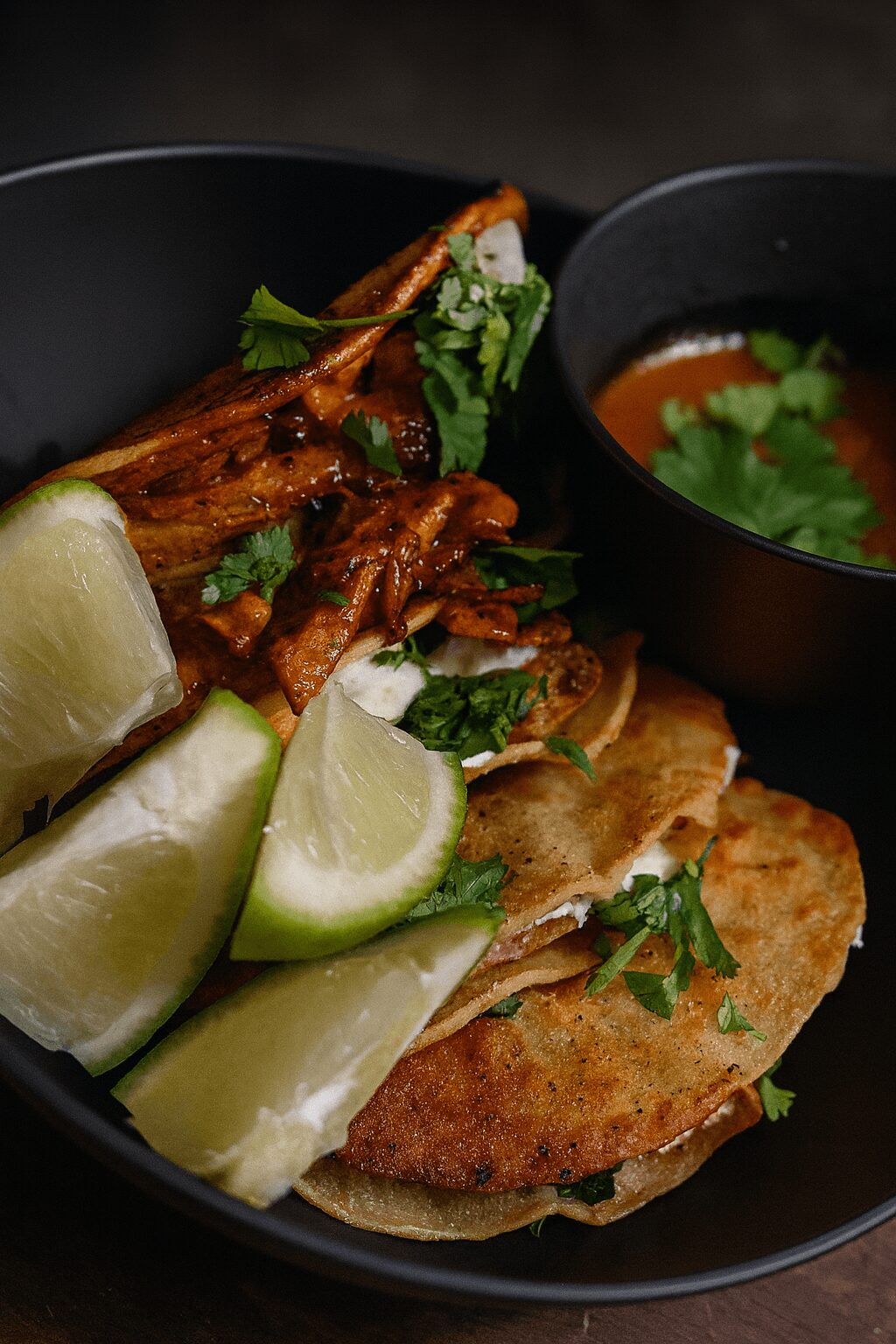 Close-up of a dish with grilled meat, lime wedges, and fresh herbs, served with a bowl of soup garnished with cilantro.