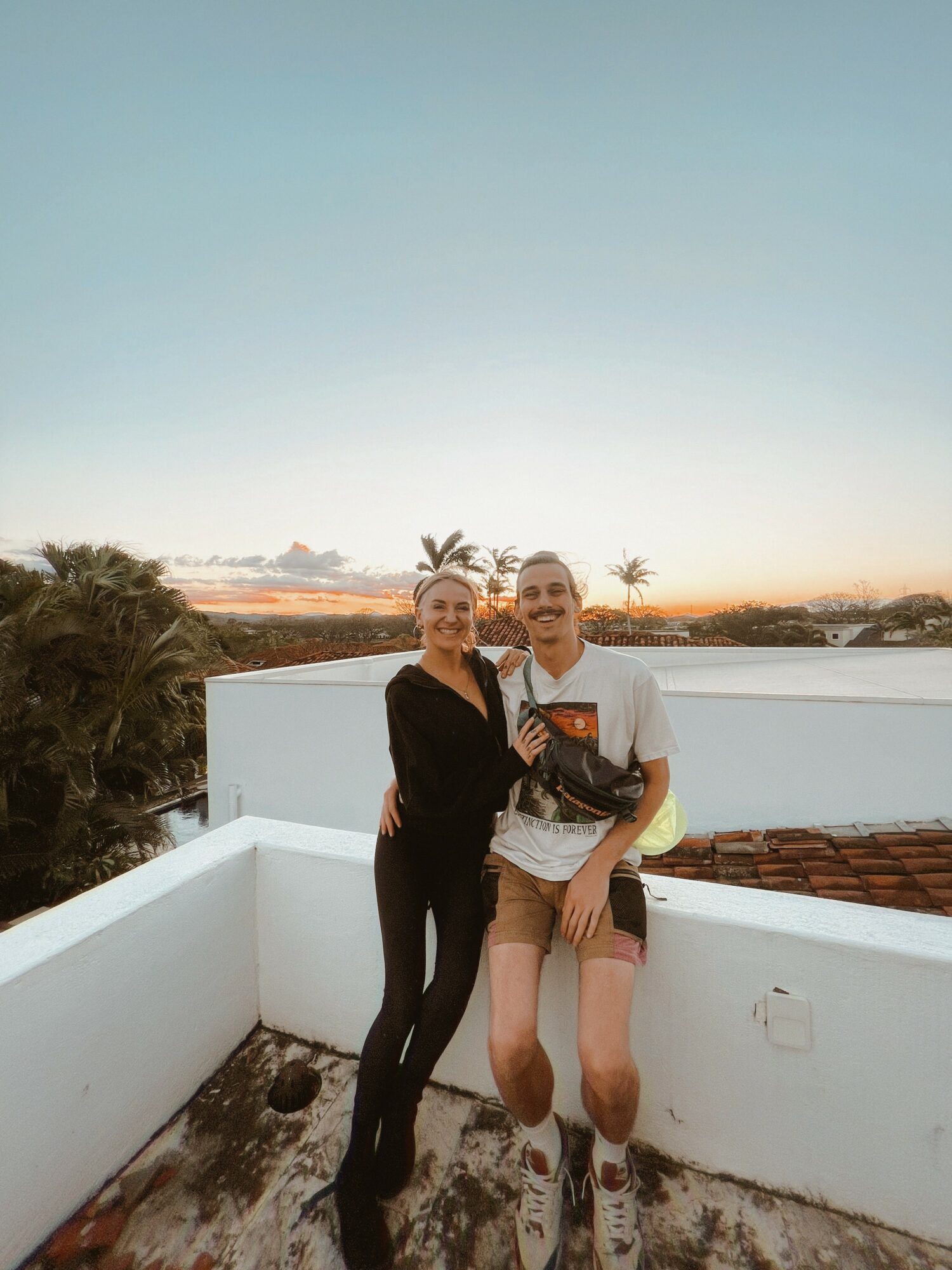 Two people standing on a rooftop with a white wall, smiling, during sunset, with trees and sky in background.