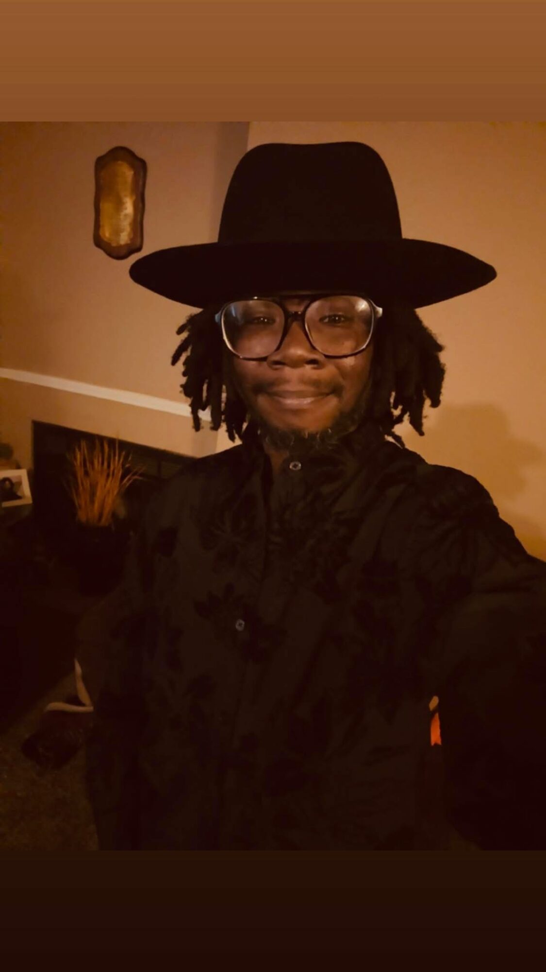 Person with glasses and dreadlocks wearing a wide-brimmed hat and black shirt, smiling indoors.
