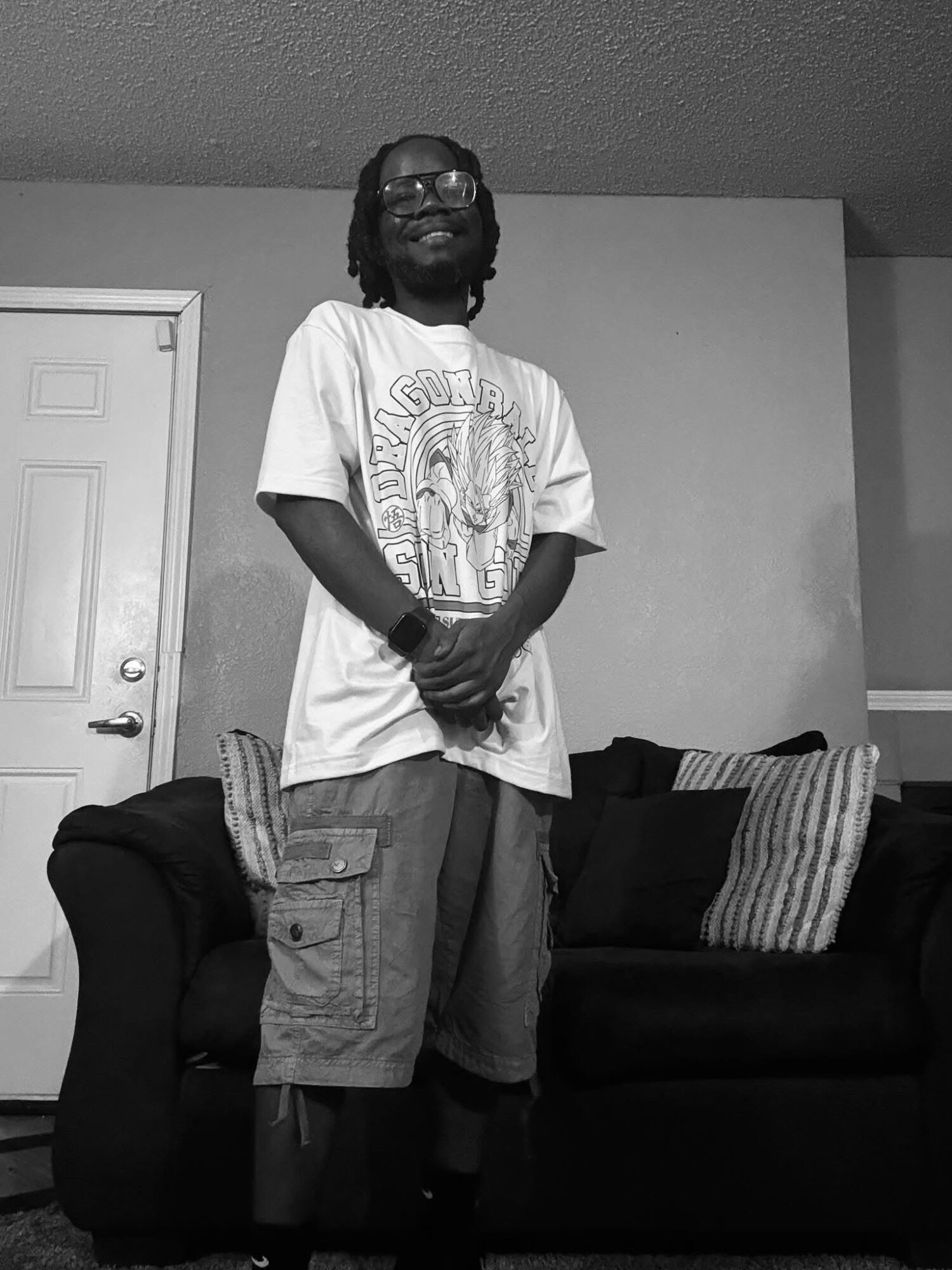 Person standing in front of a sofa, smiling, wearing a graphic t-shirt and cargo shorts, with a door in the background.