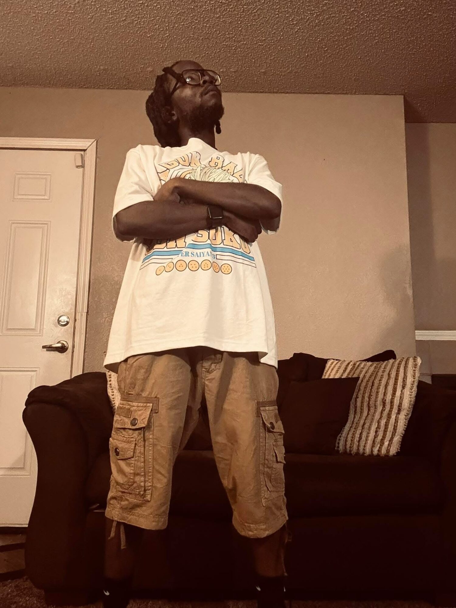 Person standing with arms crossed, wearing glasses, a white T-shirt, and cargo shorts, in a living room.