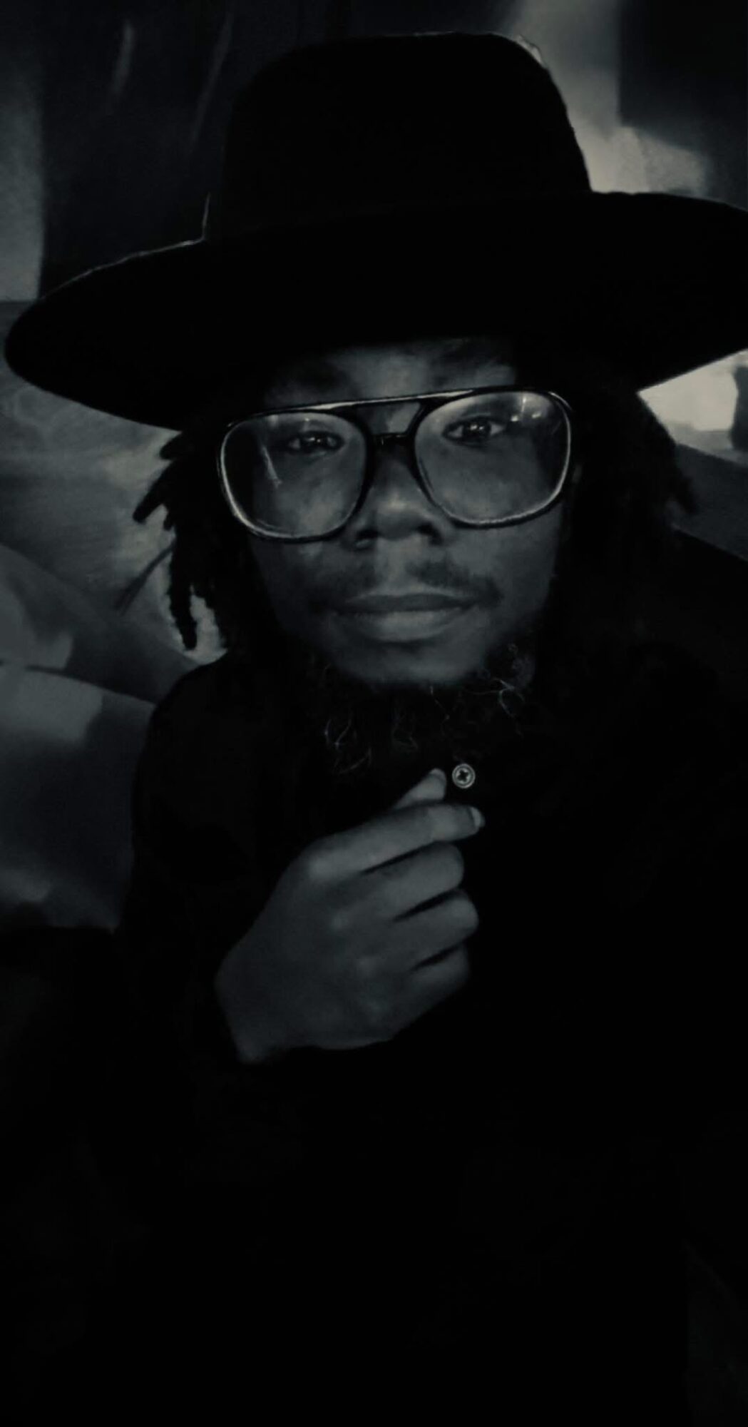 Person wearing glasses and a wide-brimmed hat, looking directly at the camera, with hand near chin, in a dark setting.
