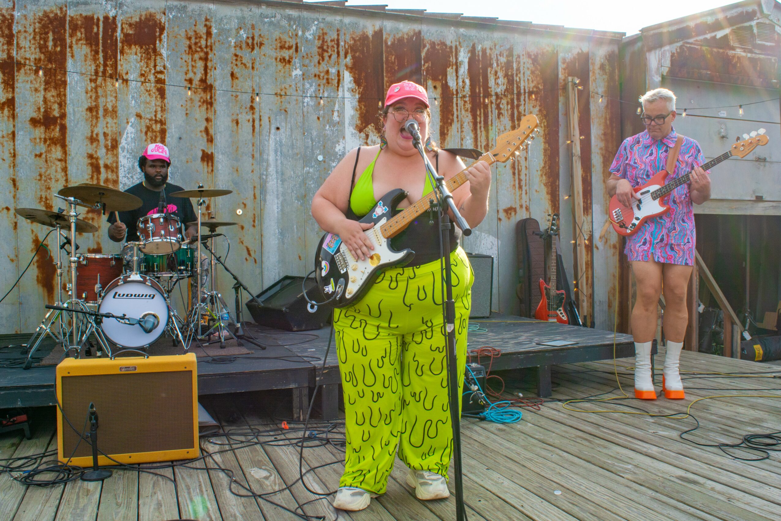 Three musicians perform on a wooden stage outdoors, with a rusty metal wall in the background. One sings and plays guitar, another drums, and a third plays bass guitar.