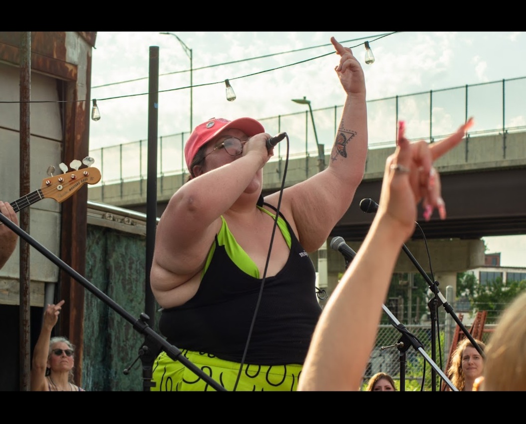 Person singing or speaking into a microphone outdoors, wearing a red hat and sunglasses, with an audience and musical instruments nearby.