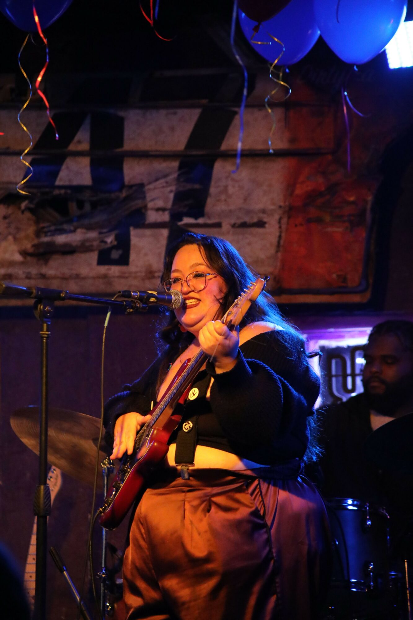 Woman with long hair and glasses smiling, holding a microphone on stage with balloons and decorations in background.