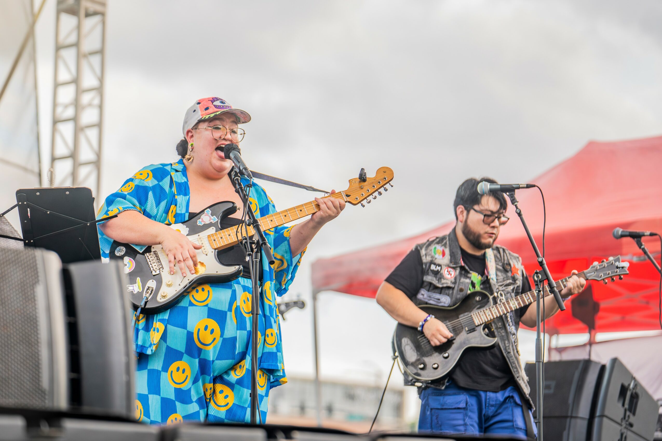 Two musicians perform outdoors under cloudy sky, one singing and playing guitar, the other playing guitar with a red tent nearby.