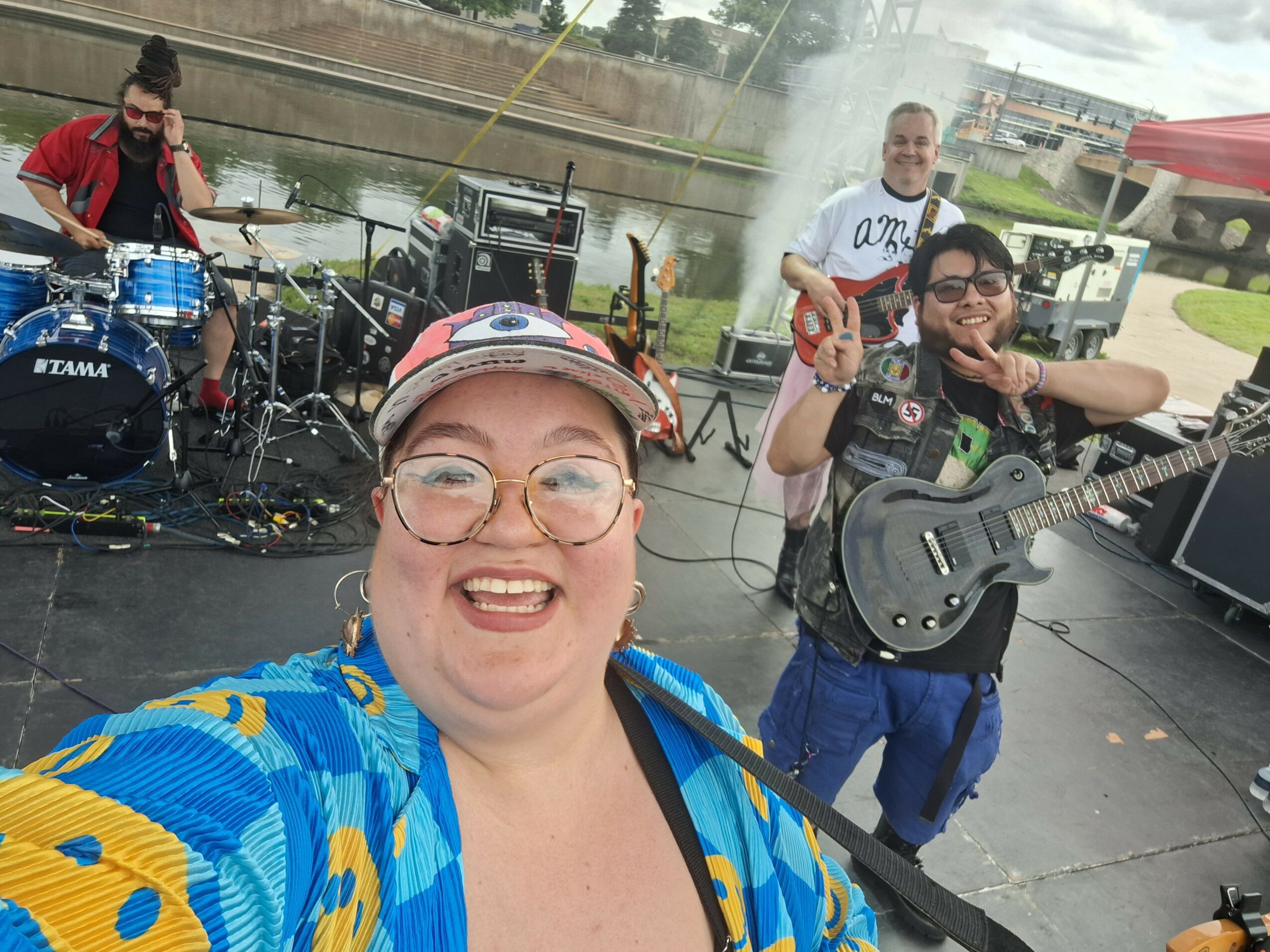 Group of four people performing music outdoors near water, with instruments and equipment, smiling and enjoying the event.
