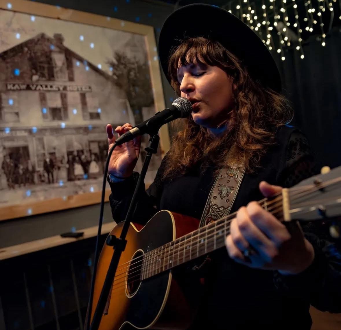Woman singing into microphone while playing guitar, wearing a hat, with a photo display and string lights behind her.