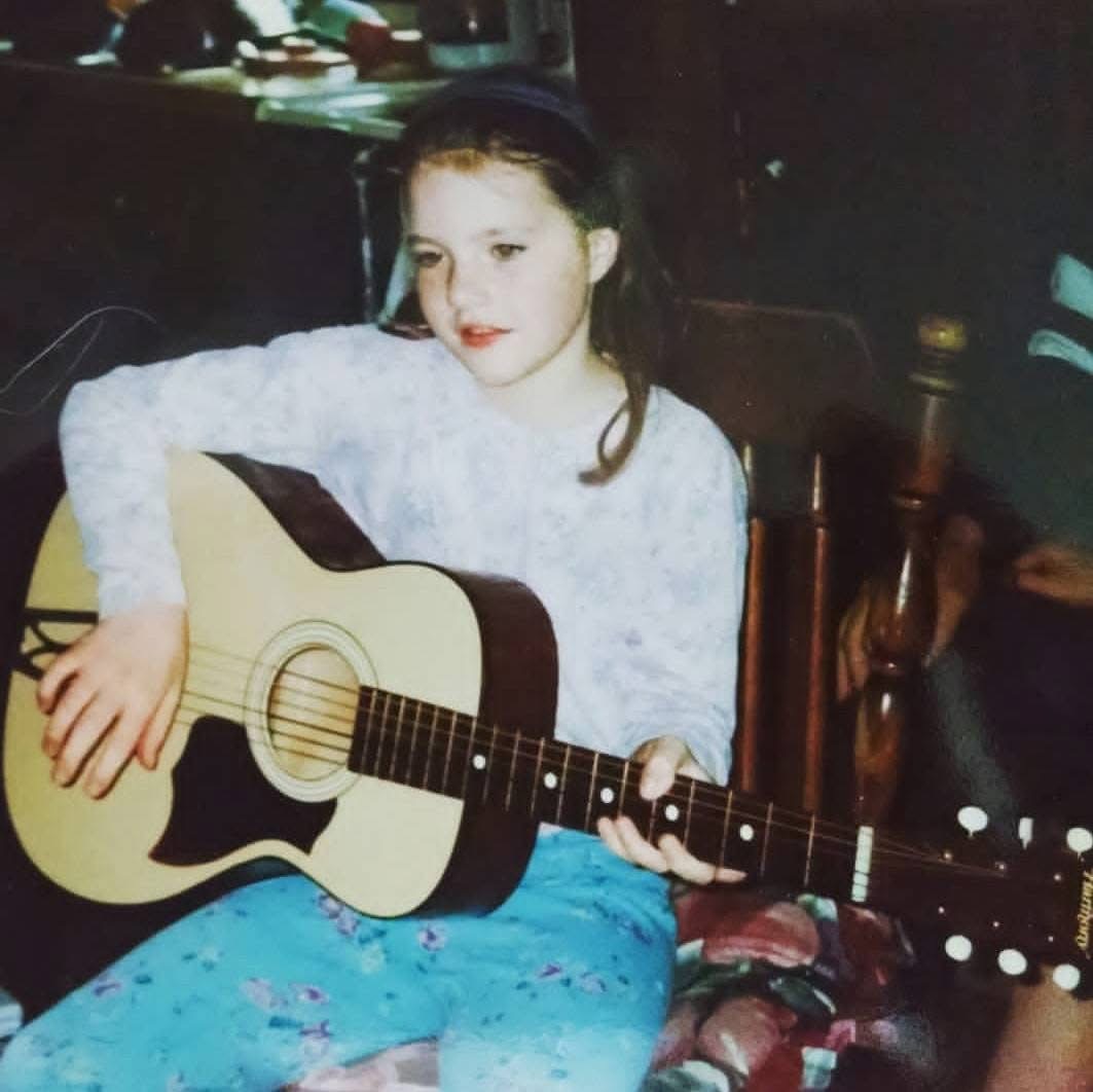 Young girl with dark hair playing an acoustic guitar indoors, wearing a white top and blue patterned pants.