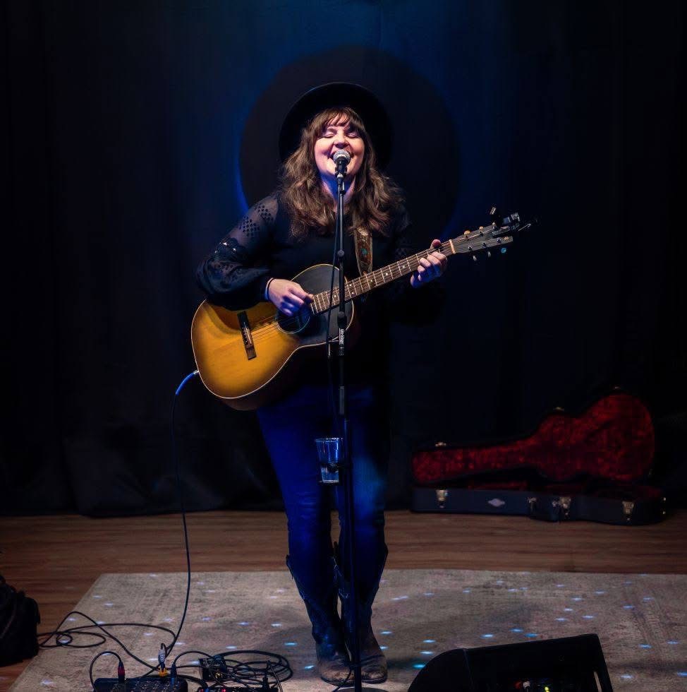 Woman with long hair, wearing a hat, playing guitar and singing into a microphone on stage.