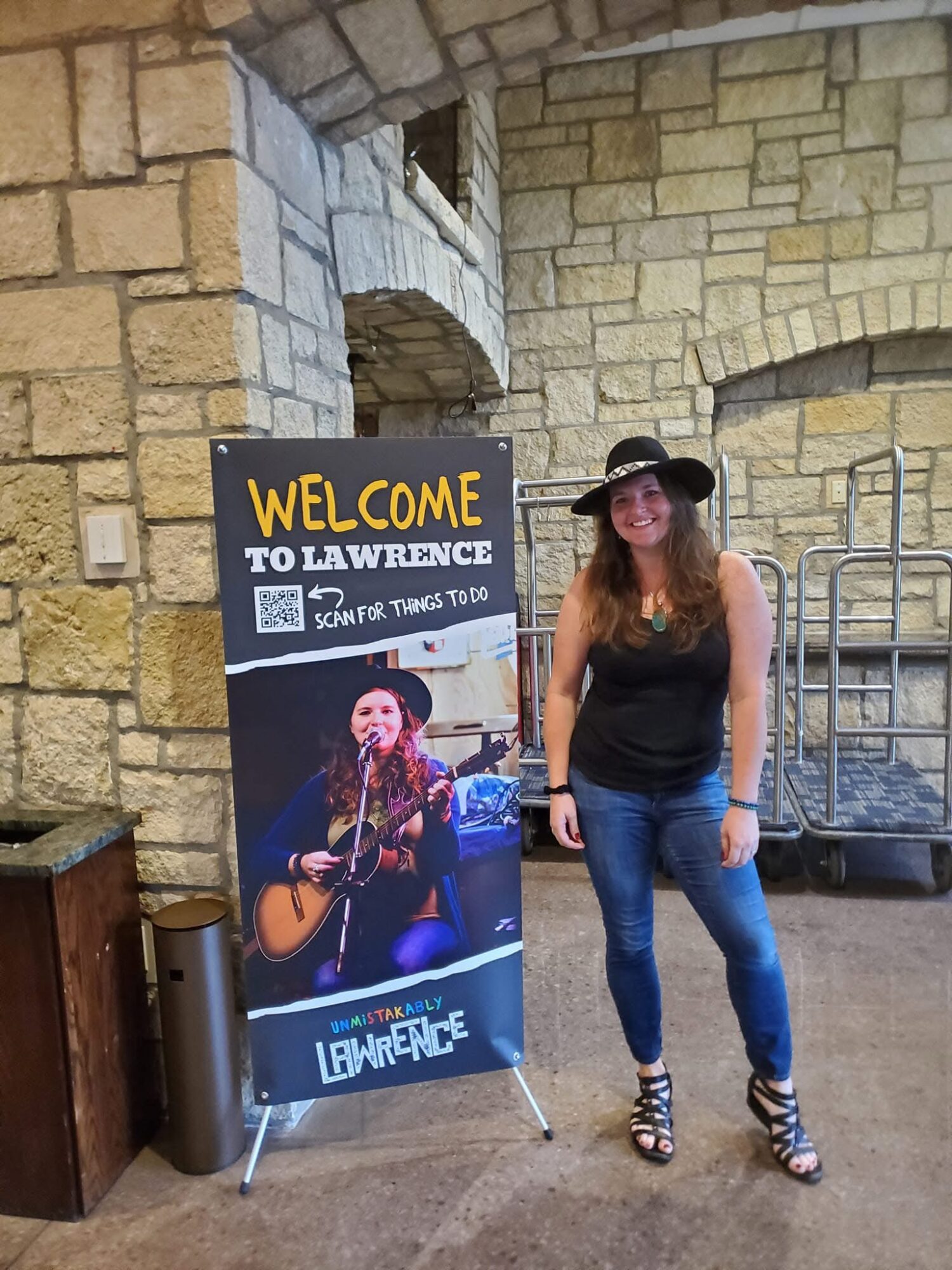 Woman with long hair, black hat, black top, and jeans standing next to a welcome sign with a musician playing guitar, brick wall background.