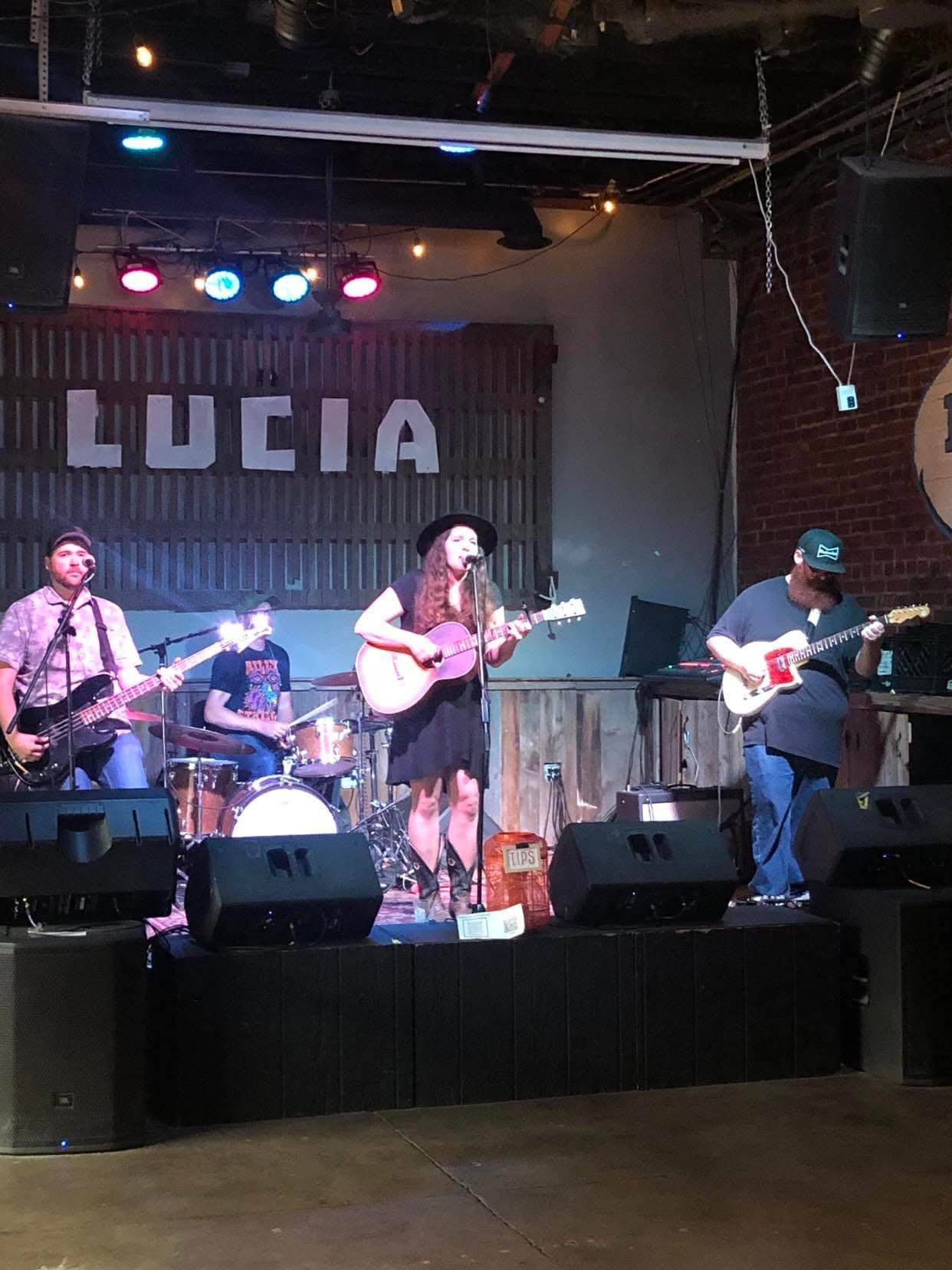 Band performing on stage with four members, one playing guitar, one singing, and others playing instruments, with a sign reading 'LUCIA' behind.