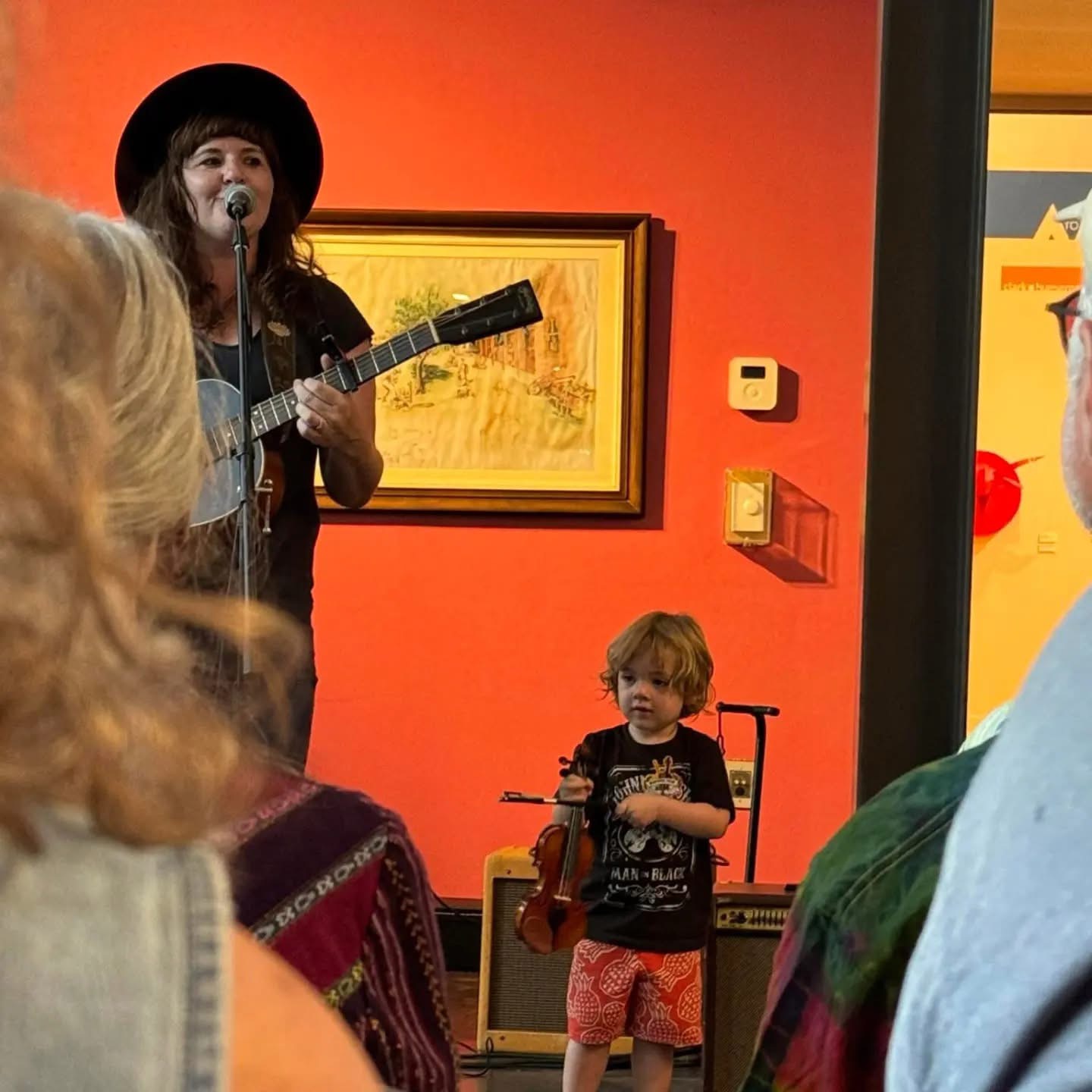 A woman with a guitar and hat performs on stage, with a young boy in front watching, in a colorful room.