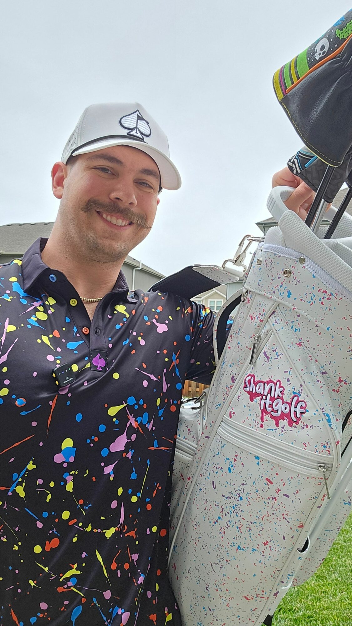 Man smiling outdoors, wearing a cap and colorful splattered jacket, standing next to a golf bag with clubs, on a cloudy day.