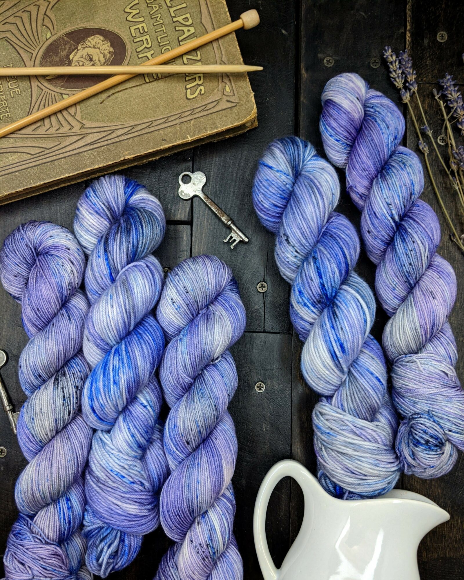 Four skeins of purple and blue yarn, a white ceramic pitcher, a vintage key, and knitting needles on a dark surface.