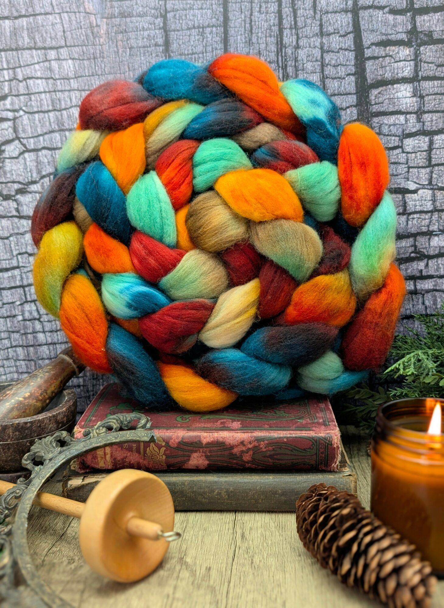 Colorful braided yarn bundle on a table with books, pine cone, and candle, against a textured wall background.