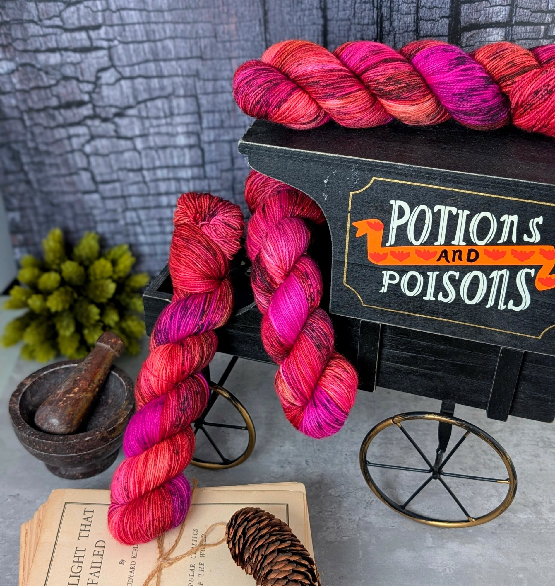 Two skeins of pink and red yarn draped over a black box with a sign reading 'Potions and Poisons,' a small plant, and a wooden pipe.