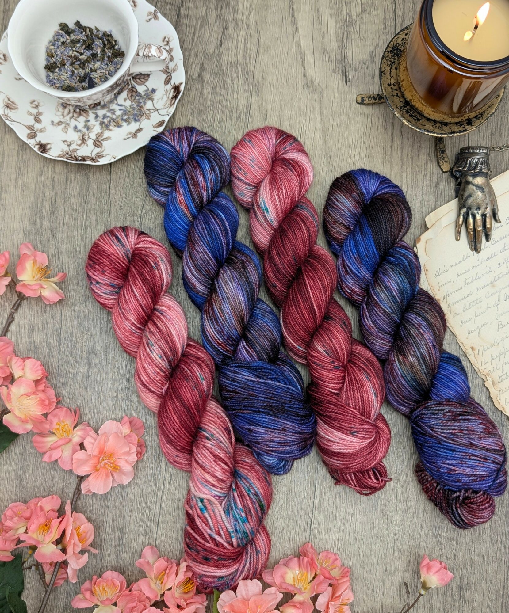 Six skeins of pink and blue variegated yarn arranged on a wooden surface with flowers, candle, and teacup nearby.
