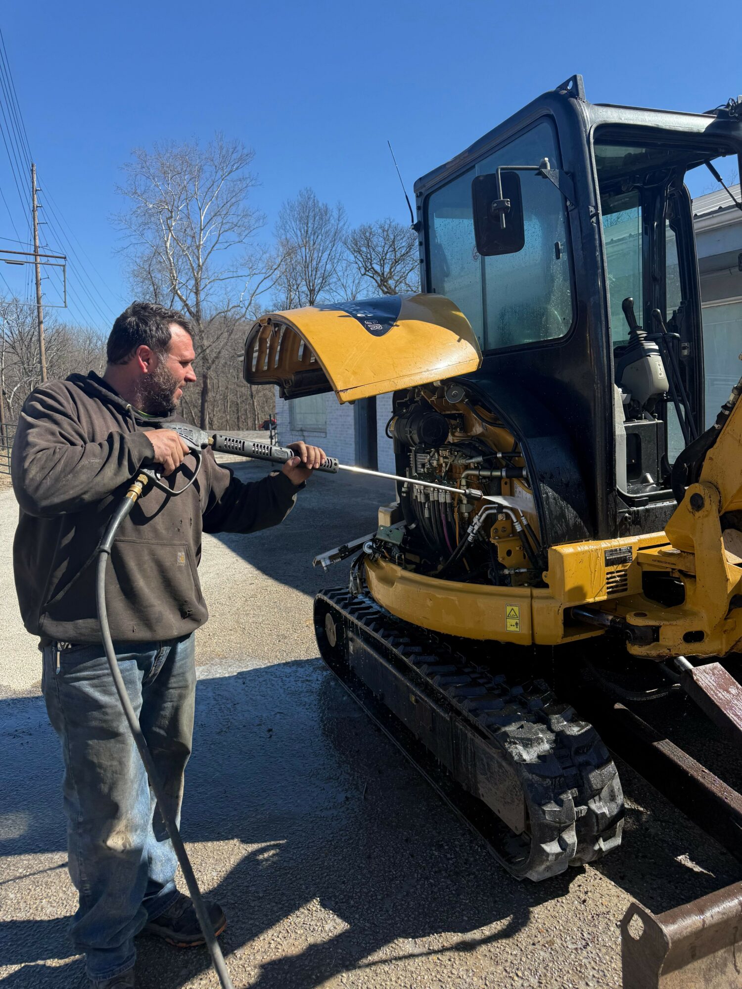 Man operating a yellow construction vehicle outdoors on a sunny day, with trees and a clear blue sky in the background.
