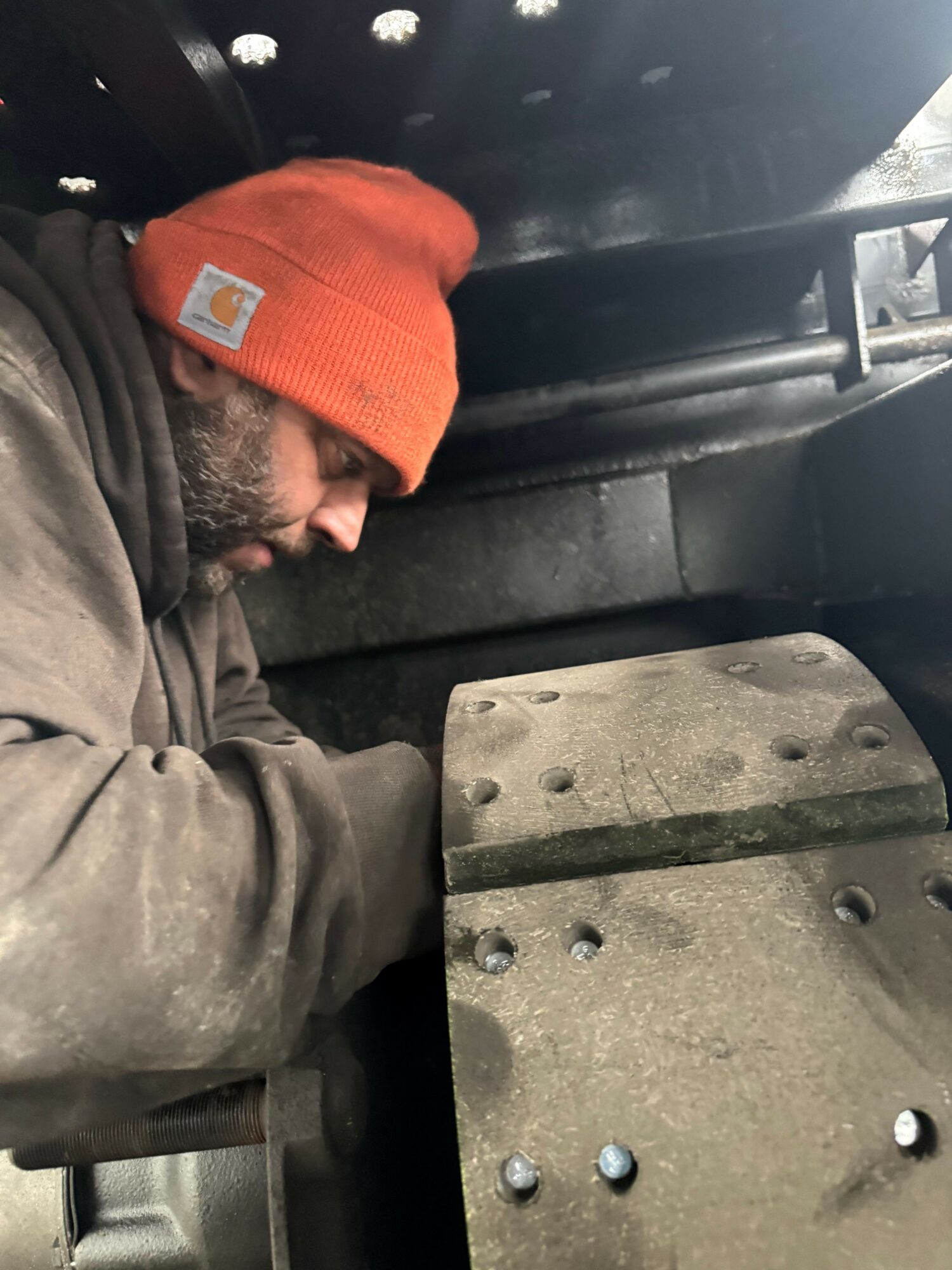 Man wearing an orange beanie and brown jacket working on machinery, with metal parts and tools visible.