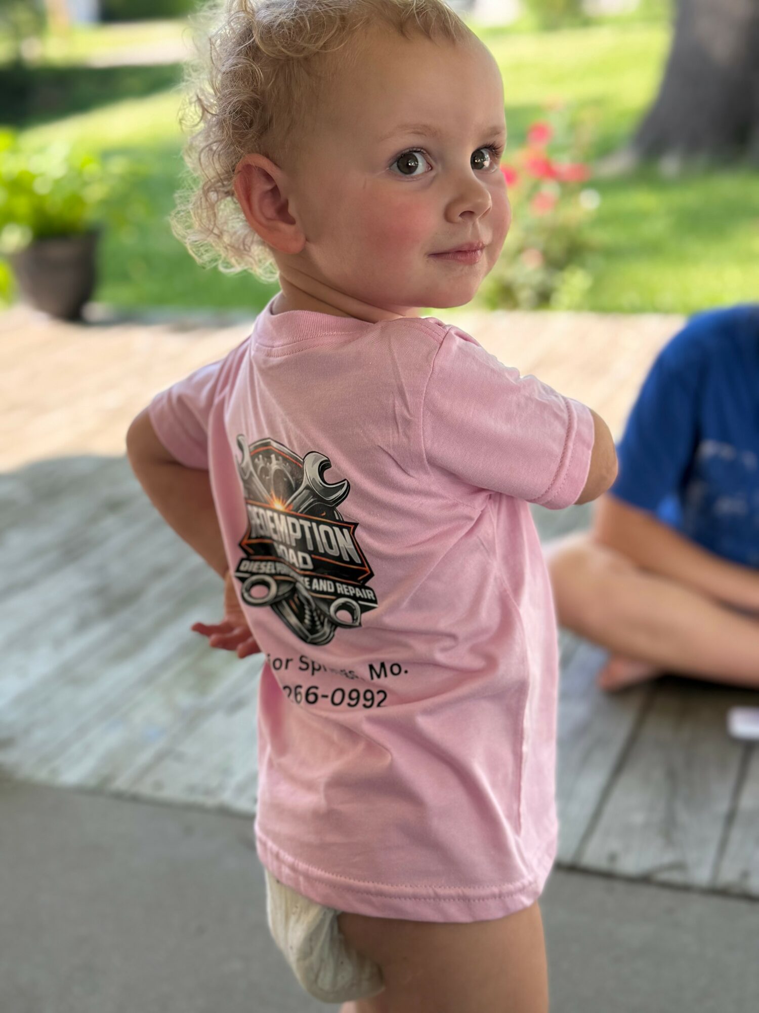 Young girl with curly hair in pink shirt outdoors, looking over shoulder, blurred background, partial view of another person, wooden surface.