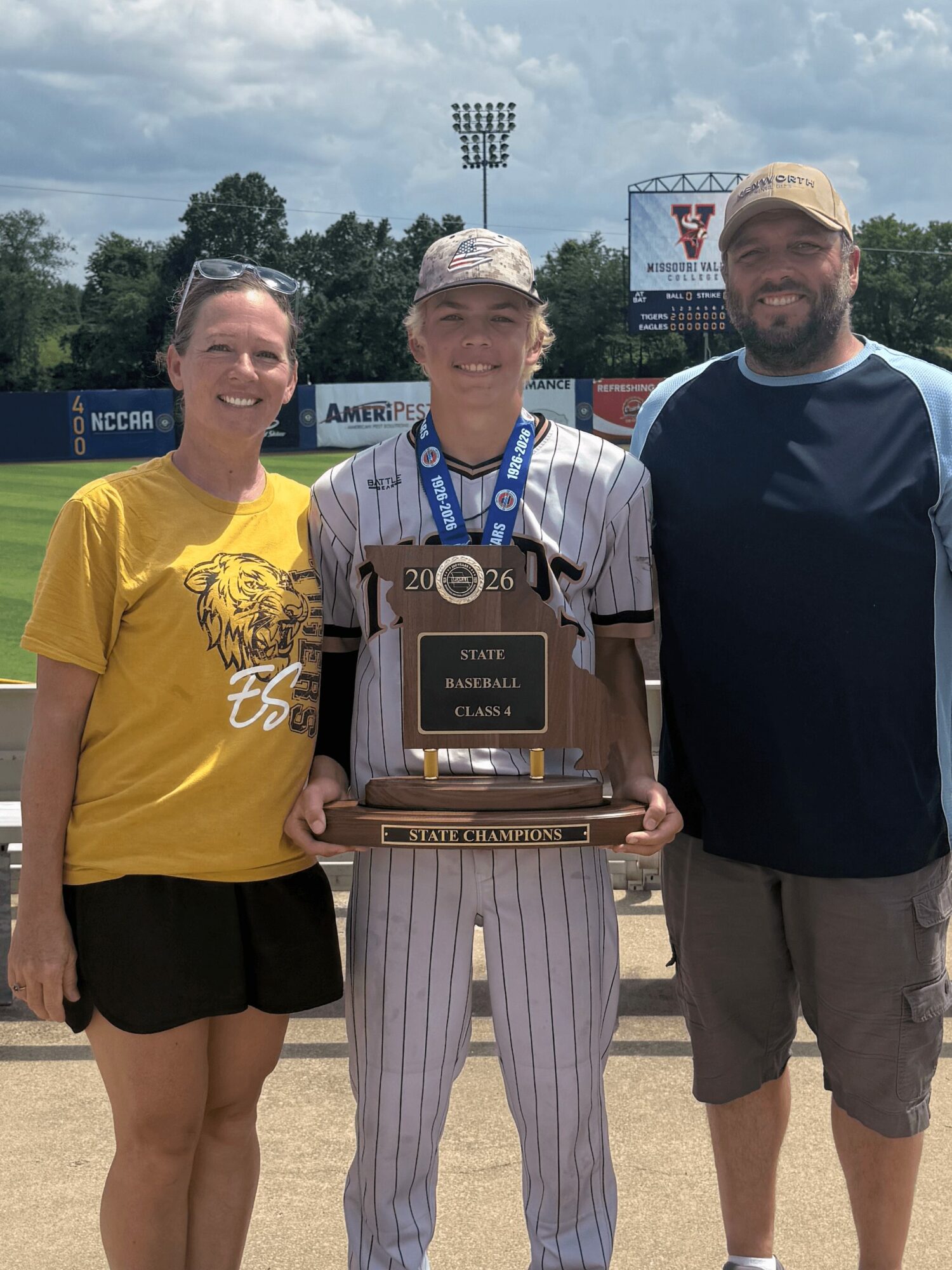 Three people standing outdoors, one holding a trophy, smiling, with a baseball field and scoreboard in the background.