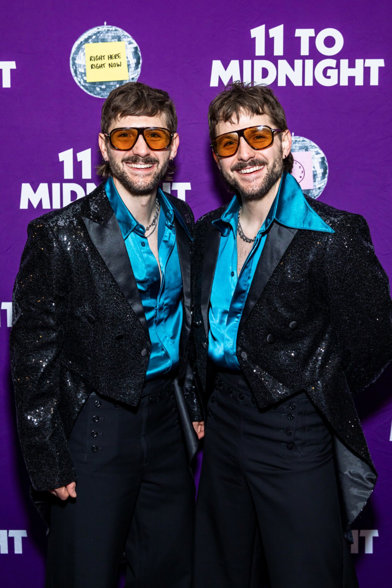 Two men in matching shiny black jackets and blue shirts stand in front of a purple background with white text.