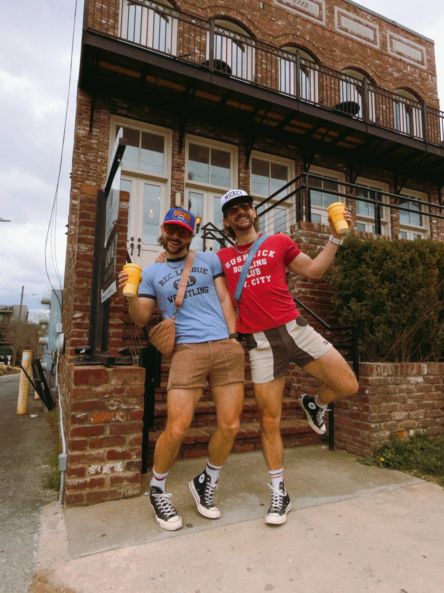 Two people in casual clothing and hats pose outdoors in front of a brick building, holding drinks and making gestures.