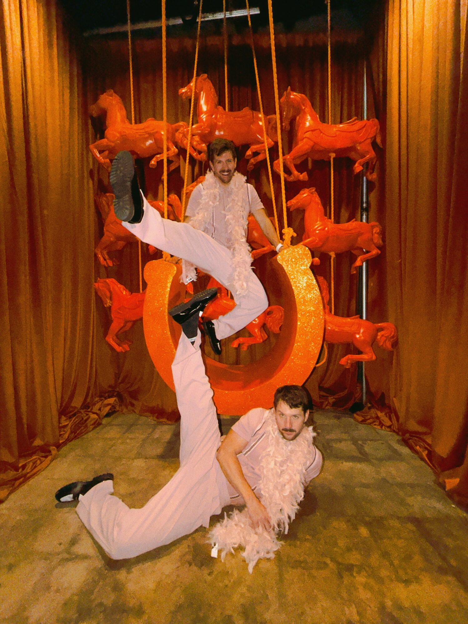 Two performers in white costumes pose on stage with a circus backdrop, one standing on the other's leg, surrounded by orange figures.