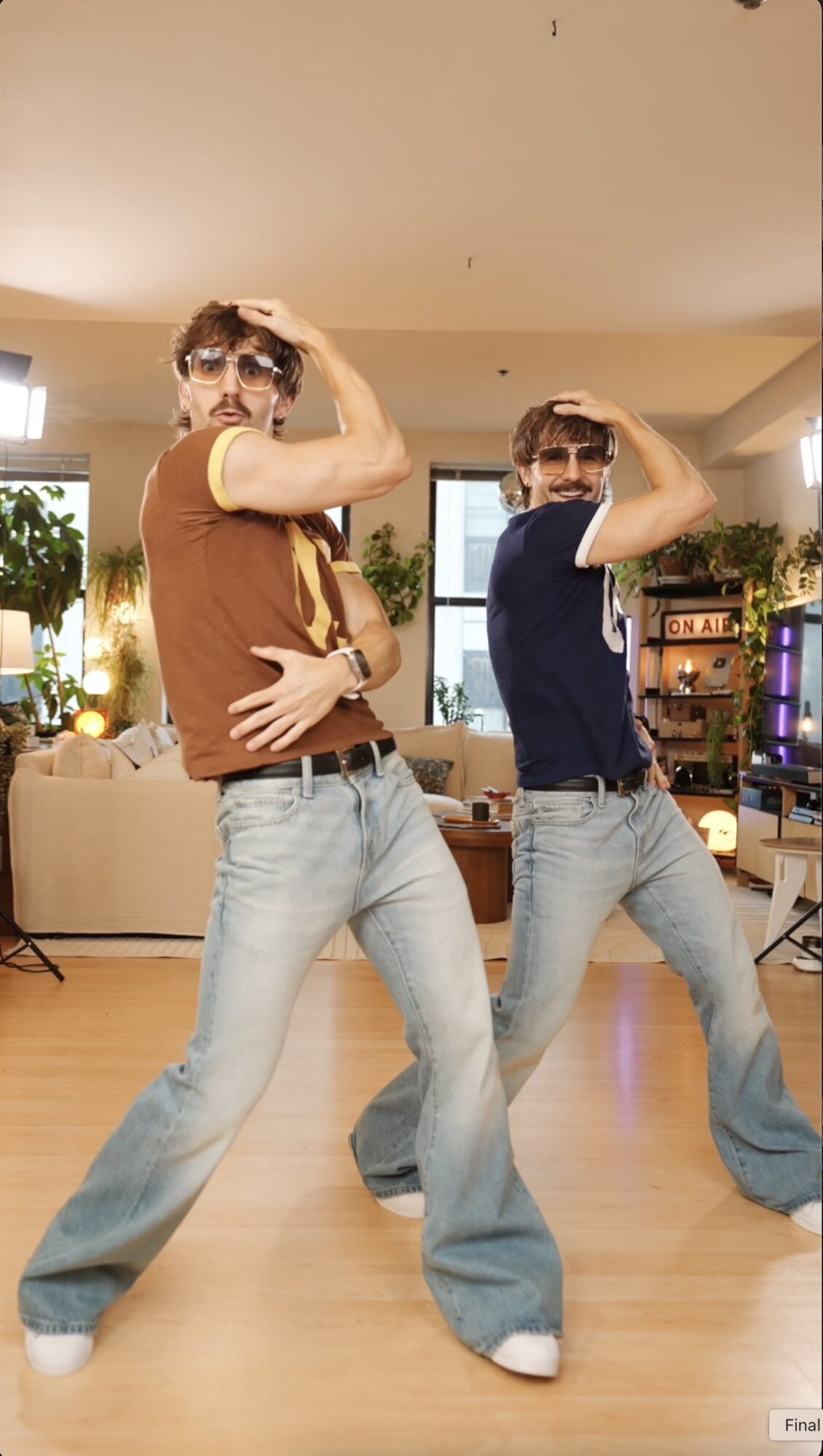 Two young men pose with arms flexed in a living room, with furniture and plants in the background.