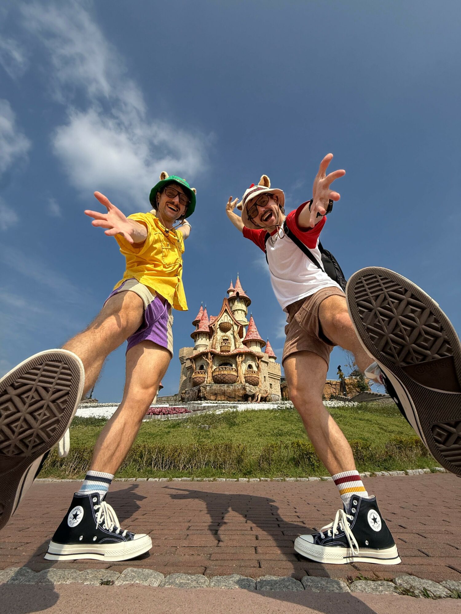 Two children jumping in front of a fountain, one in yellow and purple, the other in red and white, outdoors.