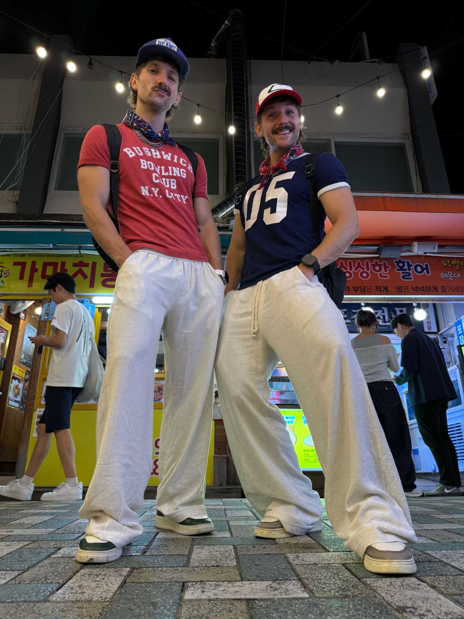 Two young men in casual clothing standing on a sidewalk at night, with a building and colorful signs in the background.